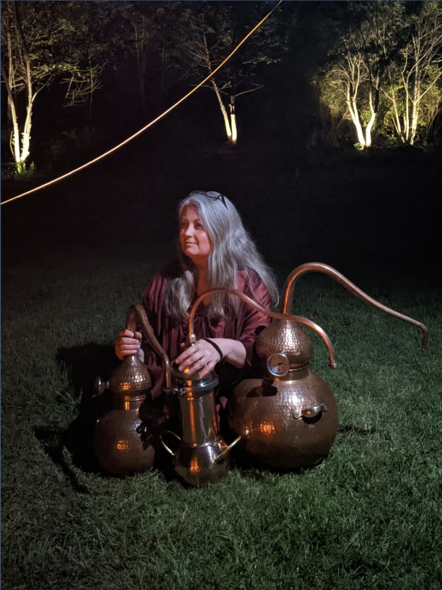 A woman with long gray hair sitting on grass at night, surrounded by copper distillation equipment, with trees illuminated in the background.