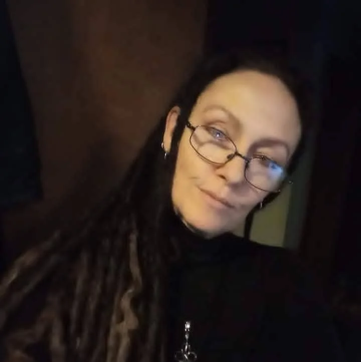 A woman with long dreadlocks wearing glasses and a black top, taking a selfie in a dimly lit environment.