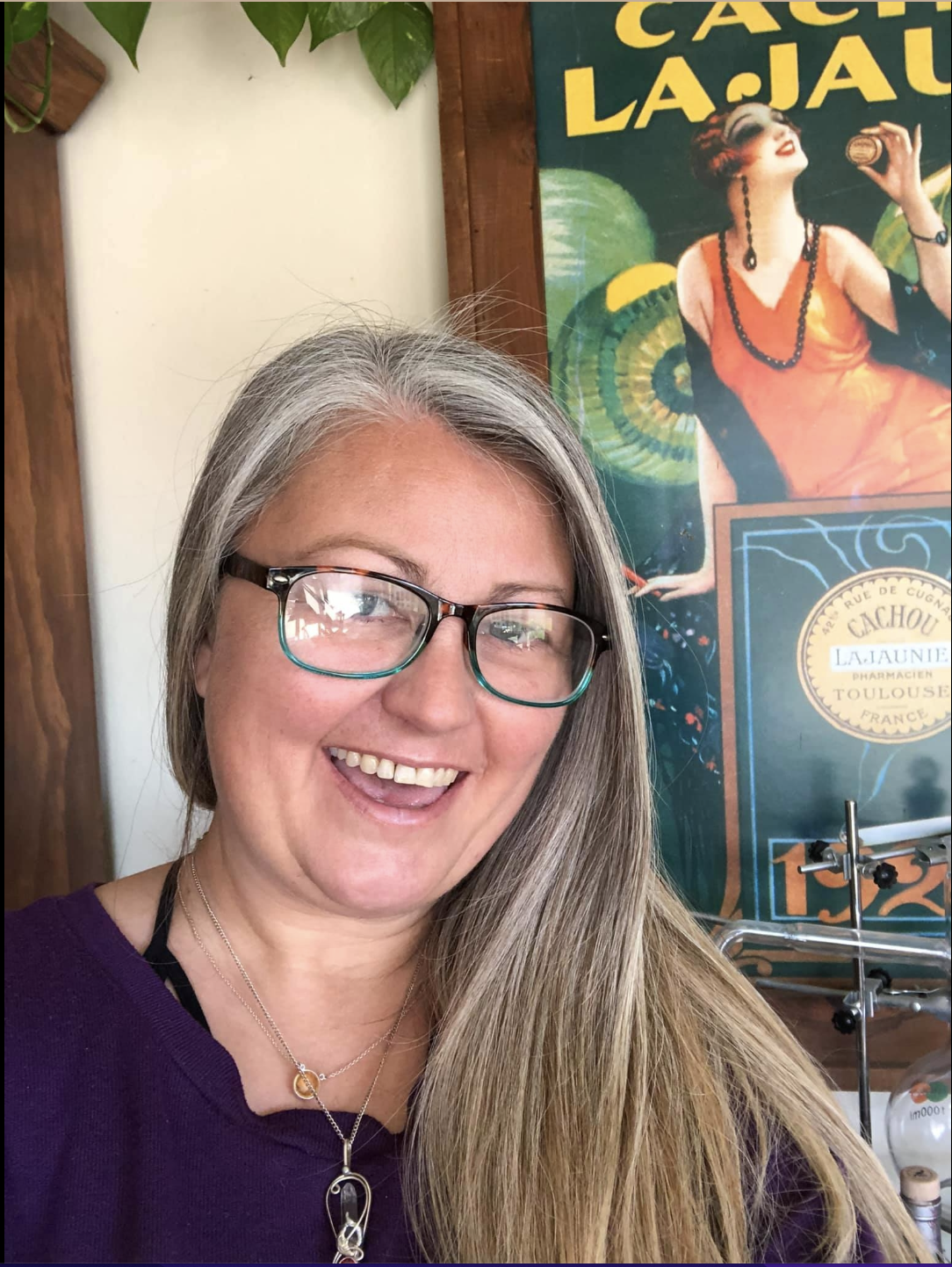 A woman with long gray hair wearing glasses and a purple top, smiling with her teeth visible, standing indoors near a colorful vintage poster featuring a woman in an orange dress and black accessories.