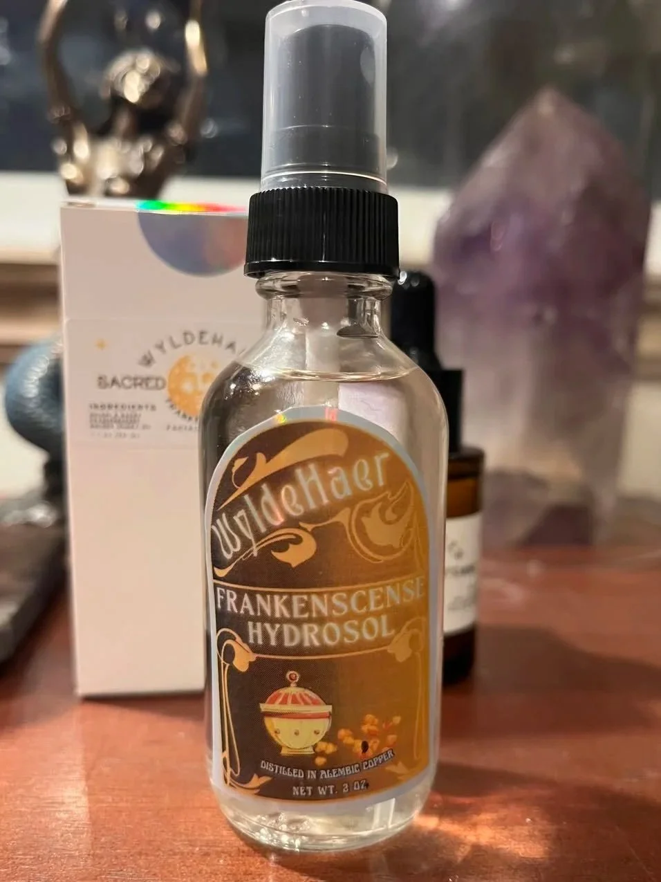 A clear spray bottle labeled 'WildeHaar Franksense Hydrosol' sitting on a wooden surface with a box and a dark bottle in the background.