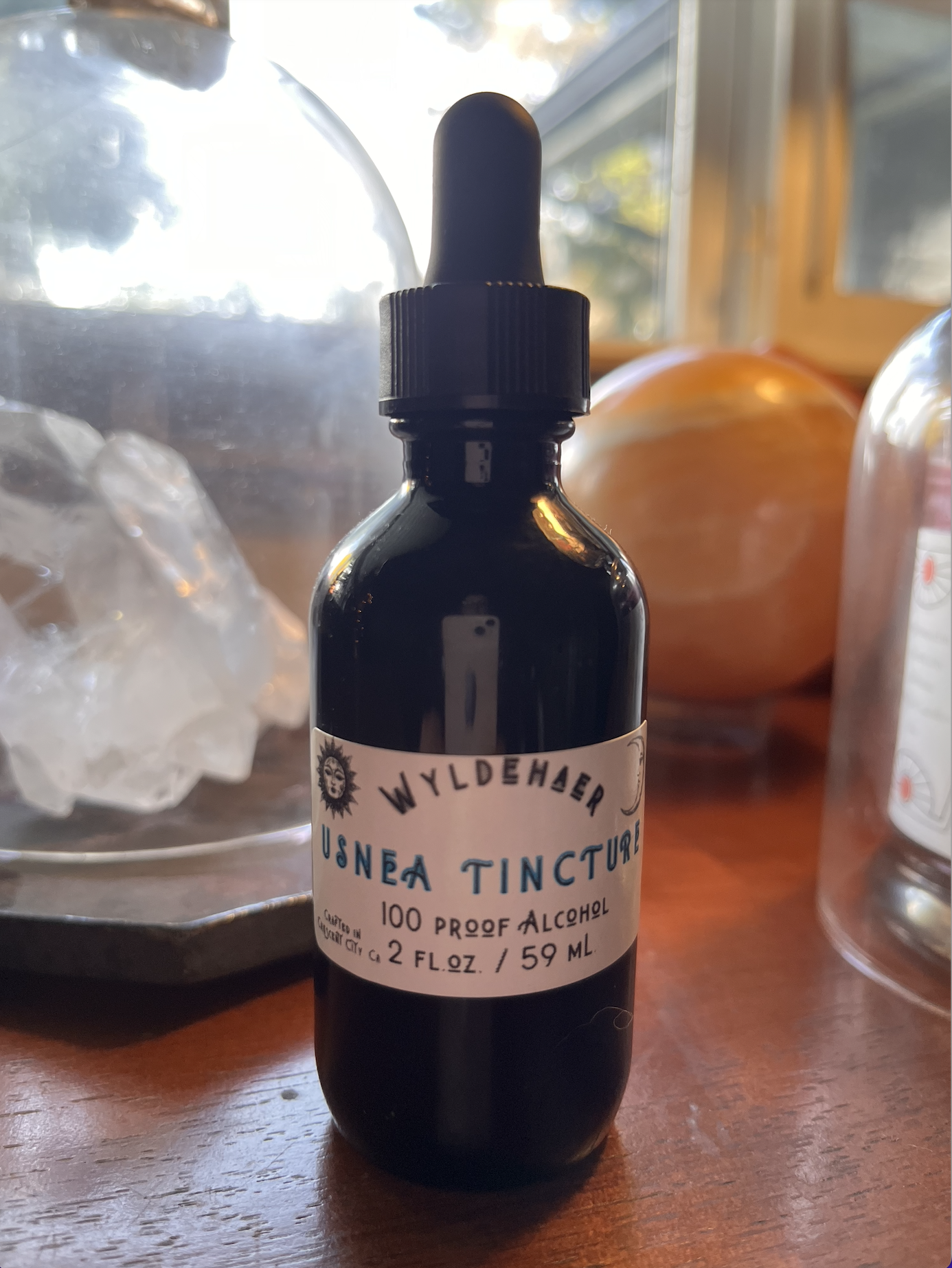A small dark glass dropper bottle labeled "Wyldehar USNEA TINCTURE," indicating it contains 100 proof alcohol and approximately 2 fluid ounces (59 milliliters). The bottle is on a wooden surface near a window, with a glass jar filled with ice and a pumpkin in the background.