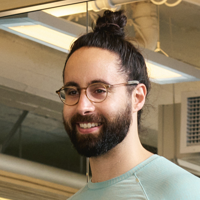 A man with glasses, a beard, and a man bun smiling indoors in a modern office or studio space.