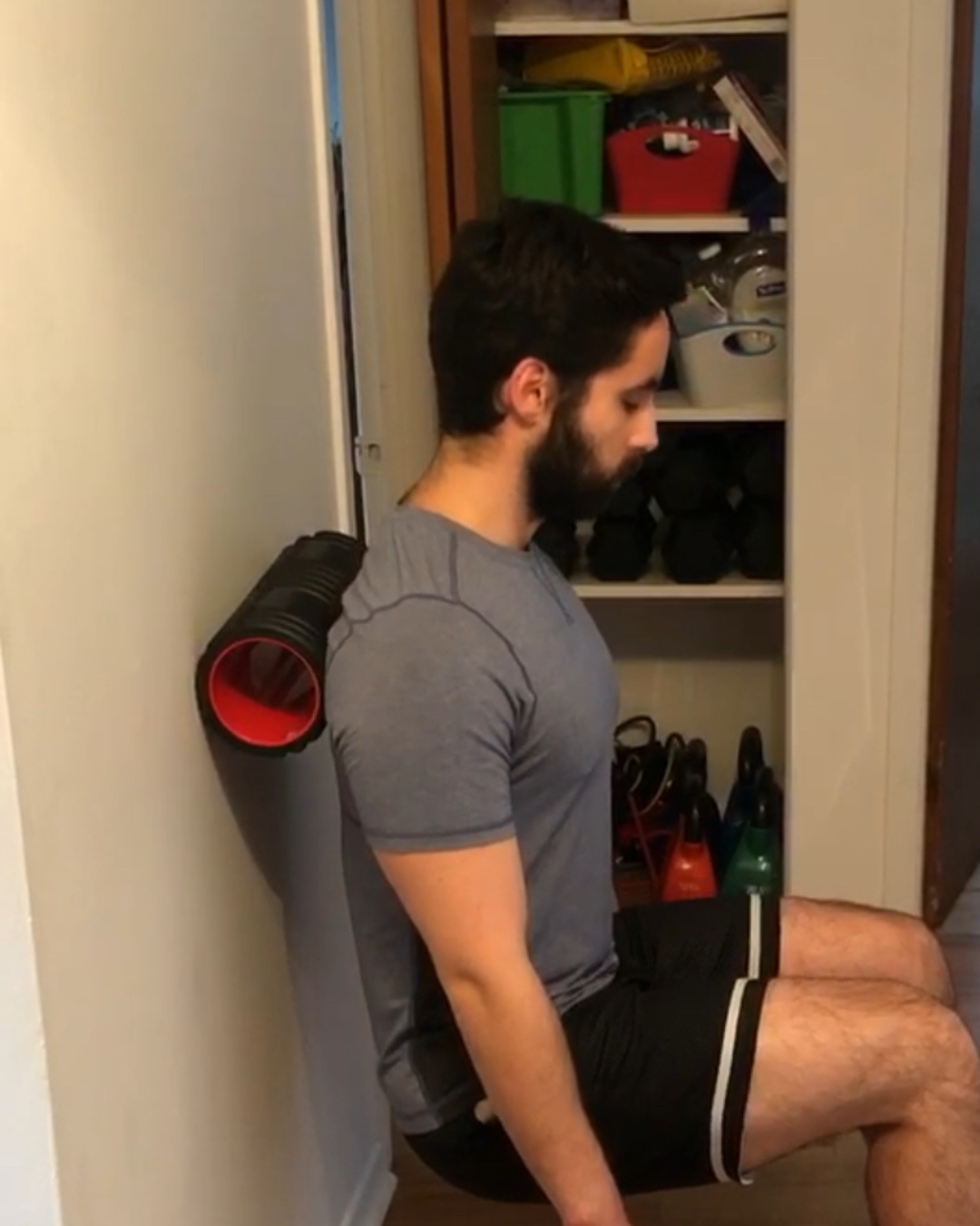 A man with dark hair and a beard sitting on the floor with his back against a wall, appearing to be resting or meditating, with a foam roller behind him and a closet filled with various items in the background.