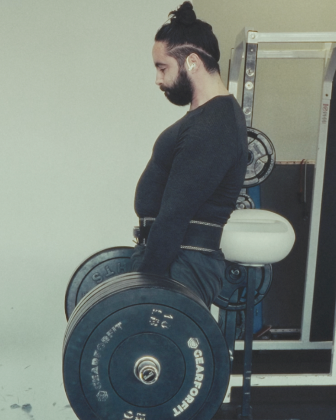 Man with a beard and long hair tied in a bun performing a deadlift exercise with a barbell at the gym.