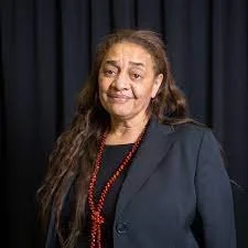 Portrait of a woman with long, wavy hair, wearing a black blazer and a red beaded necklace, standing in front of a dark curtain background.