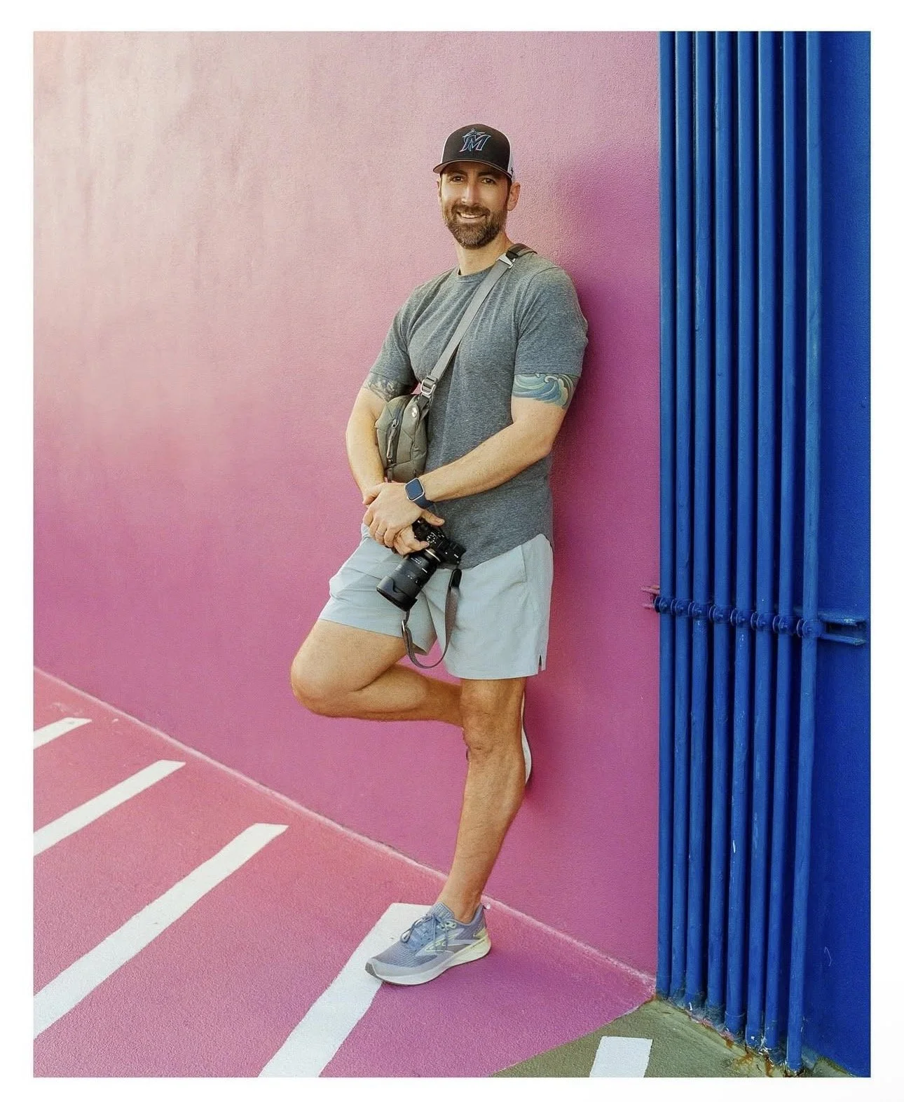 A man with a beard, wearing a black baseball cap, gray t-shirt, light gray shorts, and sneakers, leaning against a pink concrete wall with blue vertical pipes, holding a camera.