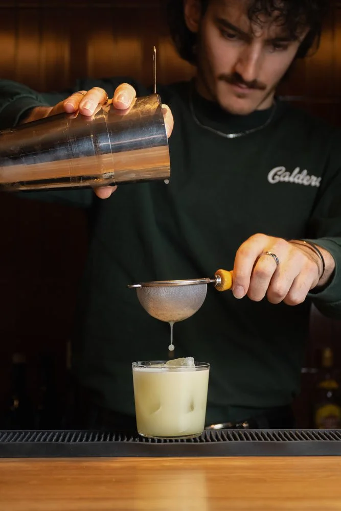 A bartender pouring a drink through a fine mesh strainer into a glass with ice, in a bar setting.