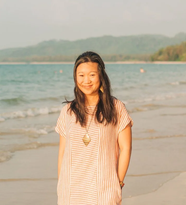 Woman smiling at the beach with water and hills in the background.