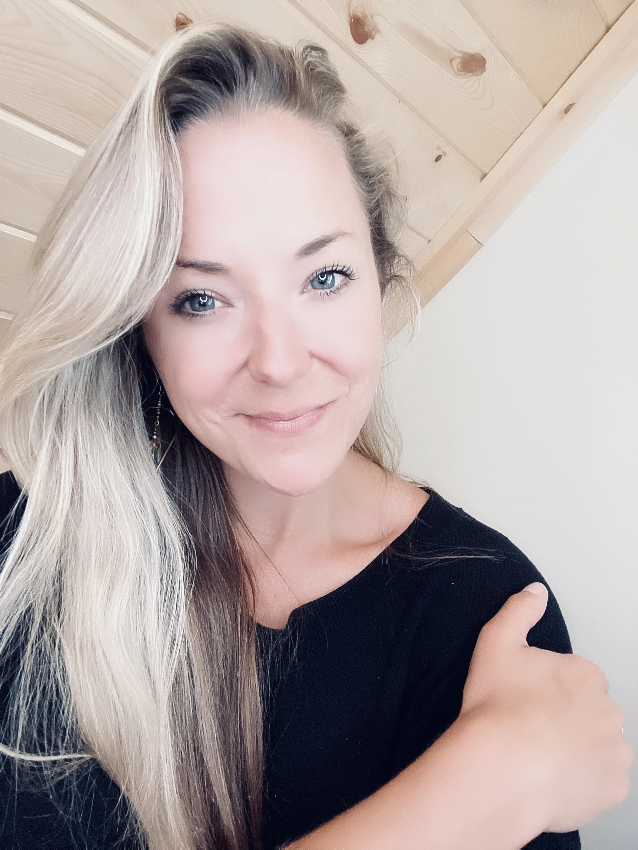 A smiling woman with long blonde hair, blue eyes, wearing a black top, standing against a wooden ceiling and a white wall.