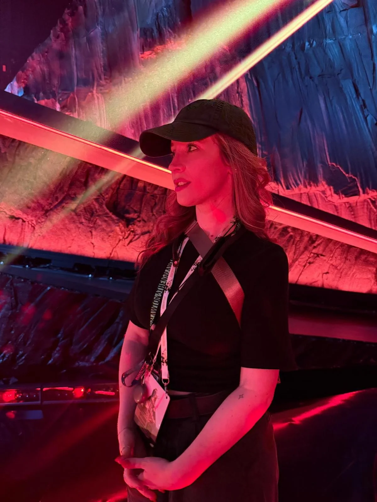 A woman with red hair wearing a black cap and black outfit, standing in front of a brightly lit, colorful, abstract background with red and pink hues.