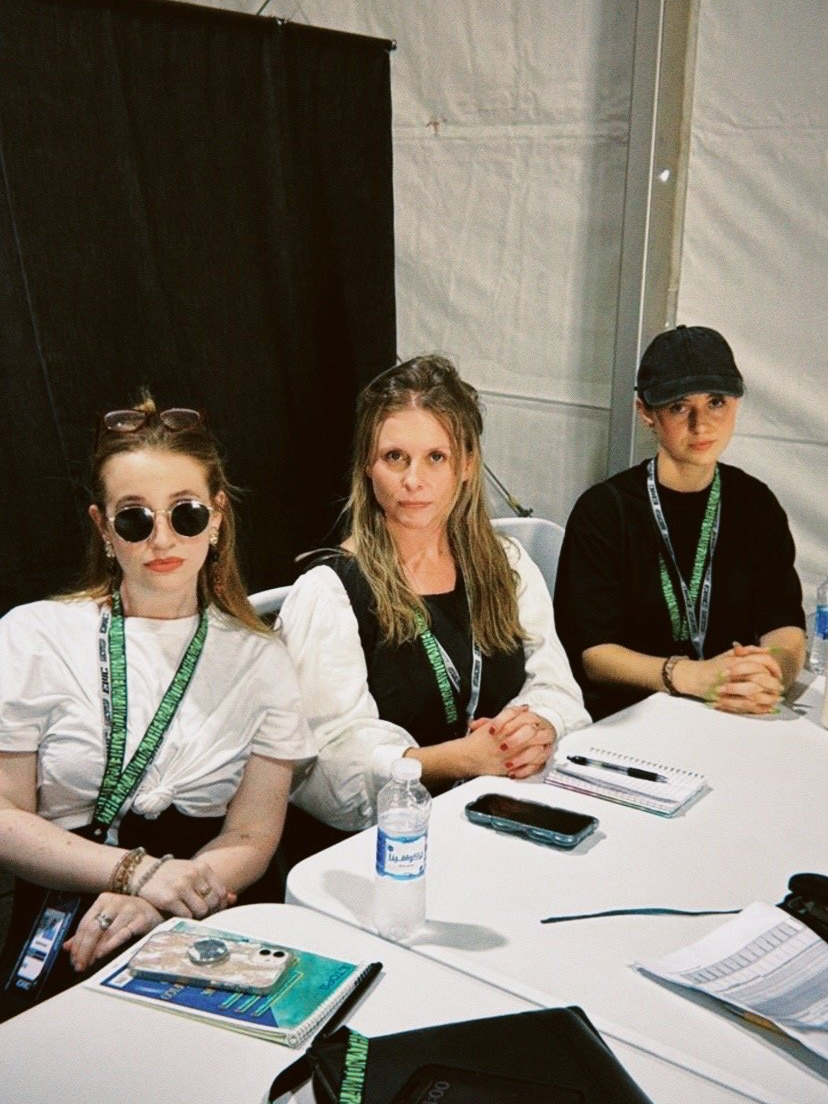 Three women sitting at a table in a conference setting, with notebooks, a bottle of water, and electronic devices in front of them.