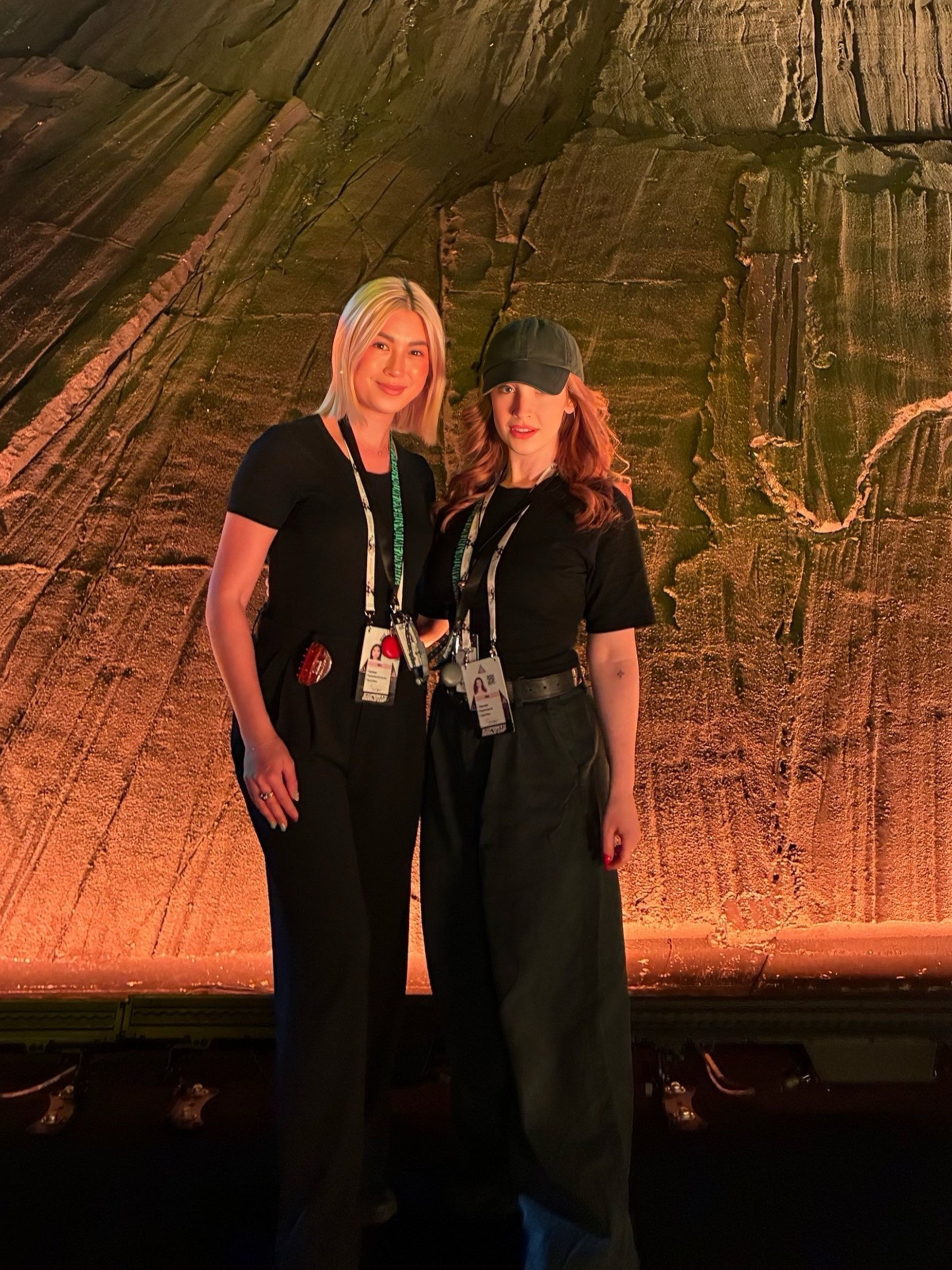 Two women standing together in front of a large rock wall, both wearing black outfits and identification badges, smiling at the camera.