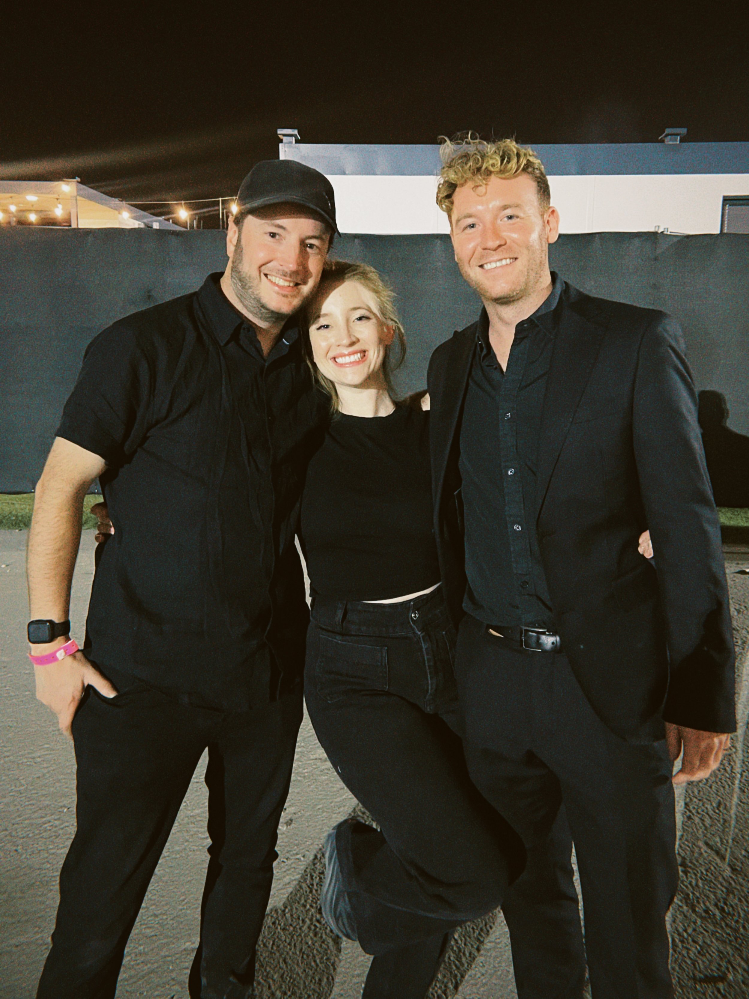 Three friends smiling and posing together outdoors at night, two men and a woman, dressed in black.