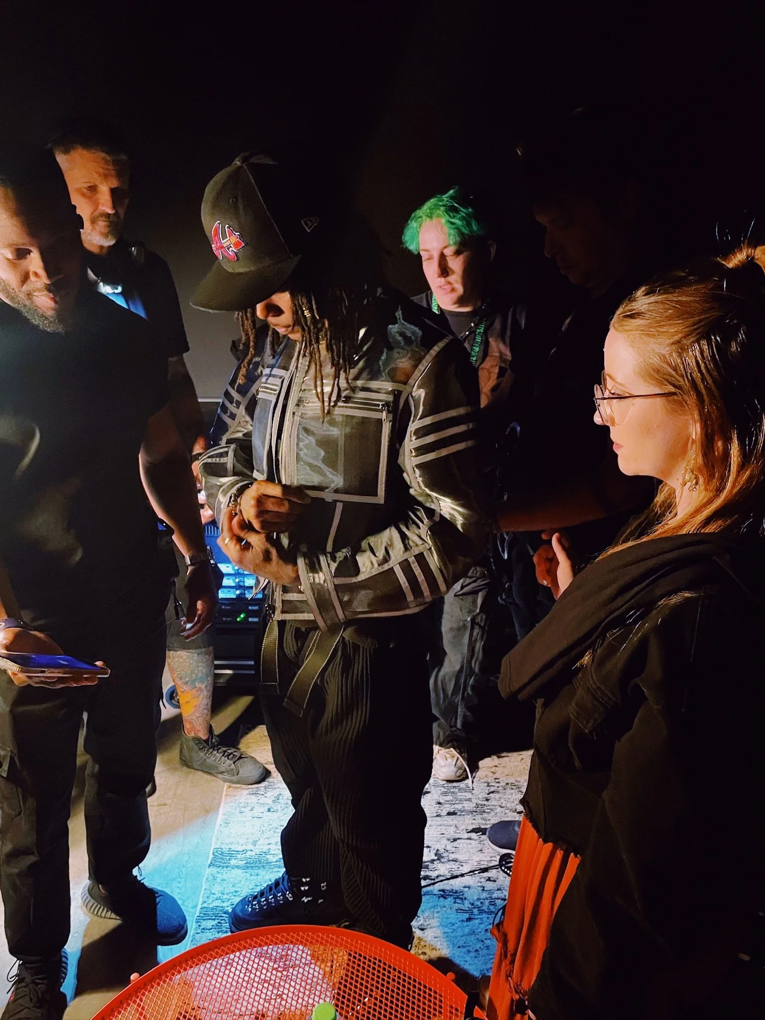 A group of people gathered closely, with some appearing to be listening intently, in a dimly lit indoor setting.