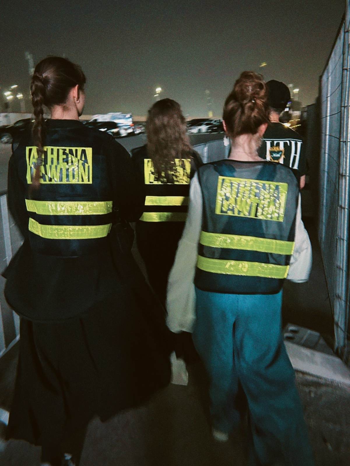 Group of women wearing reflective vests and dark jackets walking along an outdoor path at night near a parking lot.
