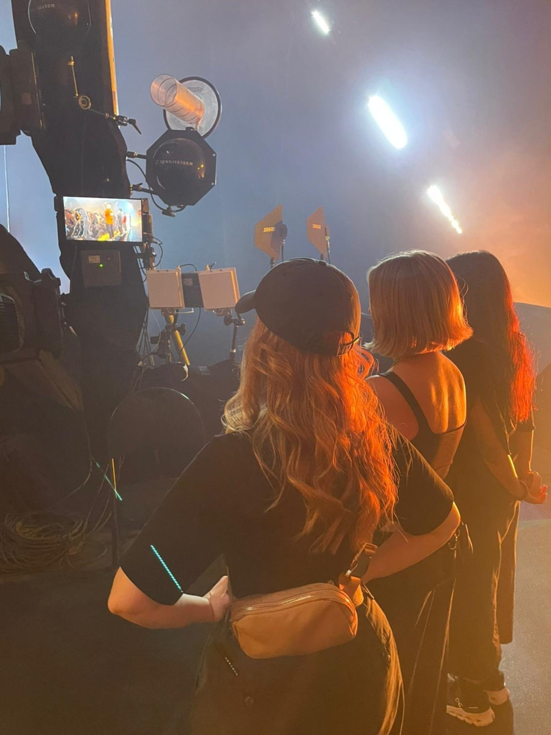 Three women standing in front of a camera and lighting equipment, watching a scene on the monitor, with bright stage lights in the background.
