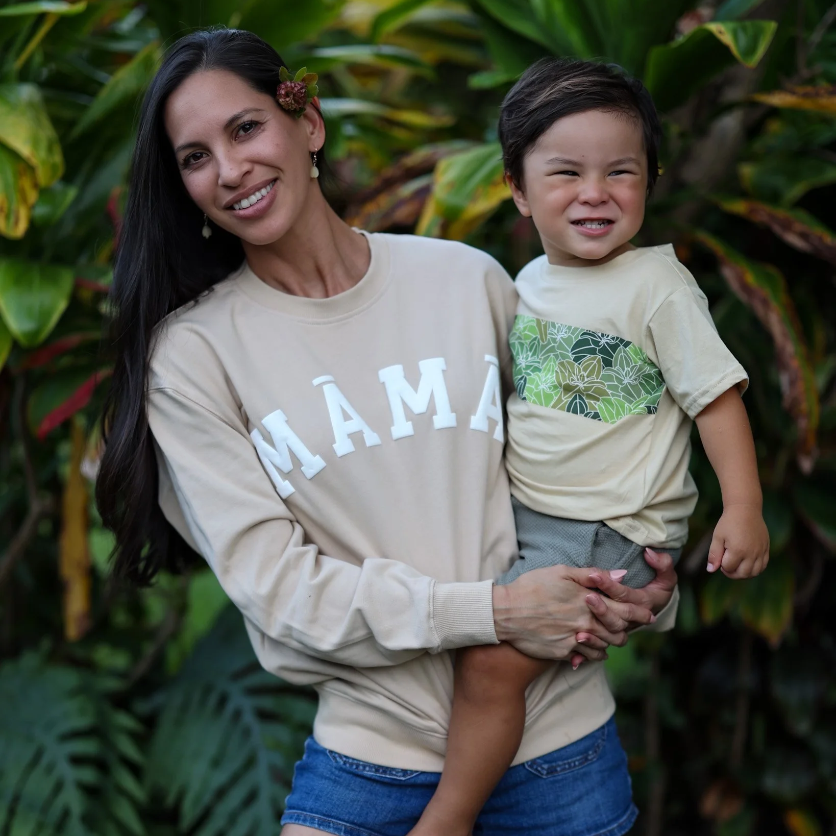 A woman wearing a beige sweatshirt with the word 'MAMA' on it is holding a young boy. The woman has long dark hair and is smiling, with a flower in her hair. The boy has short dark hair and is making a playful grimace. They are outdoors with lush gre