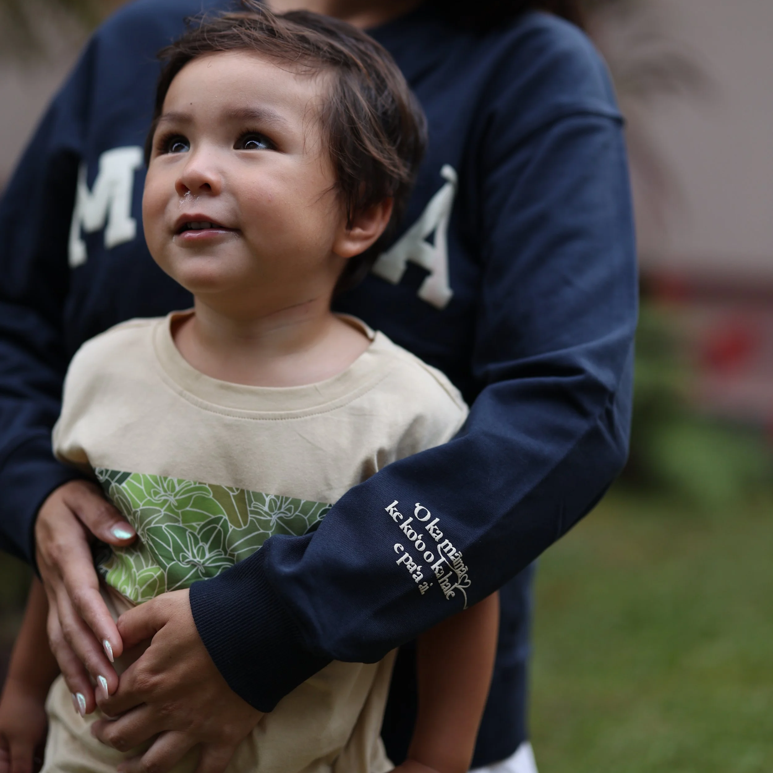 A young boy being held by an adult outdoors; the boy is looking upward with a smile, and the adult's arms are wrapped around him.