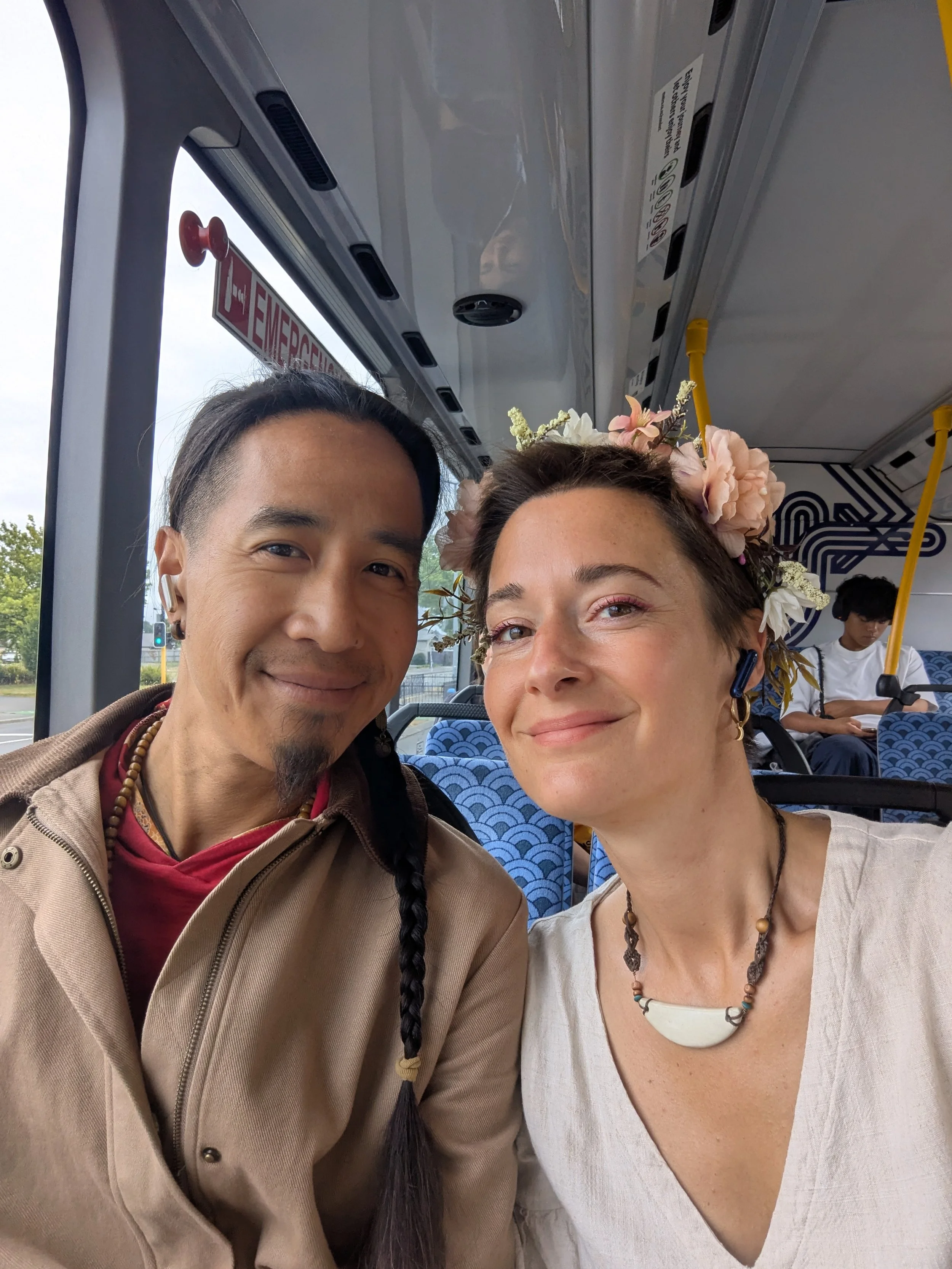 Two people sitting inside a bus, smiling at the camera. The person on the left has dark hair in a braid, and the person on the right wears a flower crown and a white top. In the background, another person is sitting and using a phone.