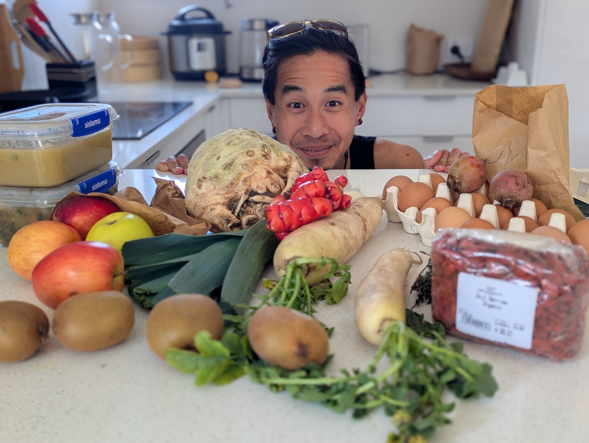 A man peeking over a kitchen counter surrounded by various fresh groceries including apples, kiwis, leeks, yams, eggs, a container of prepared food, and other vegetables.