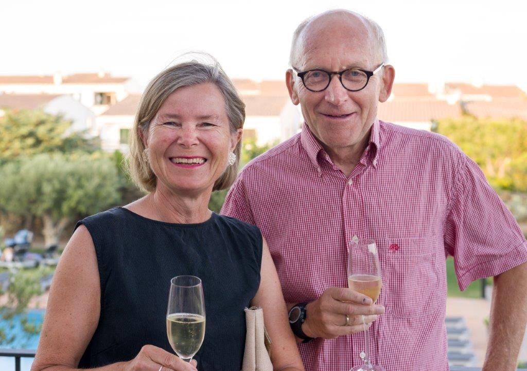 En eldre kvinne og en eldre mann smiler og holder glass med champagne på en utendørs fest, med et boligområde i bakgrunnen.