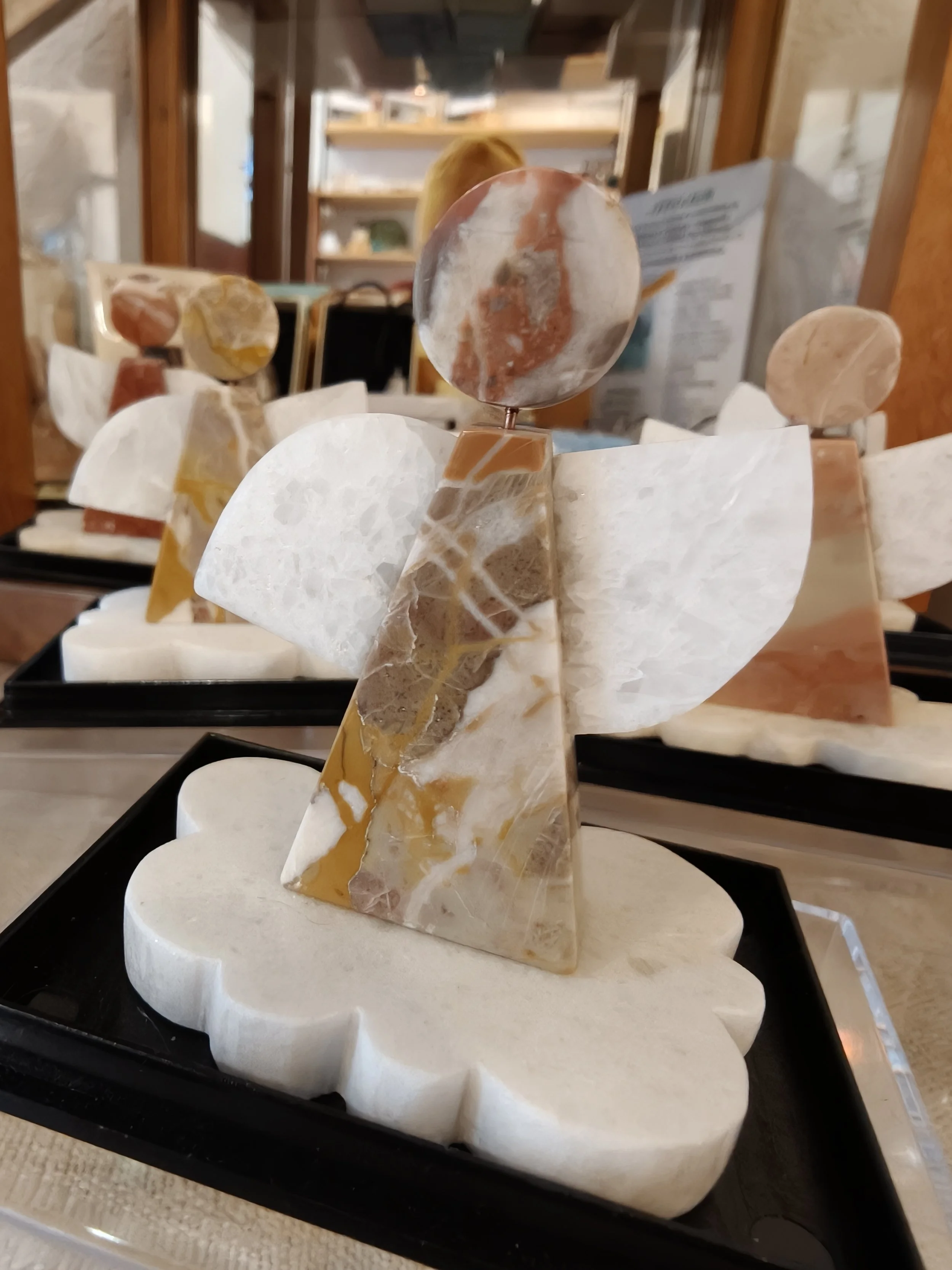 Scultura di un angelo in marmo di Arzo con diverse forme geometriche colorate, posizionata su una base bianca, in vendita in un negozio di artigianato.