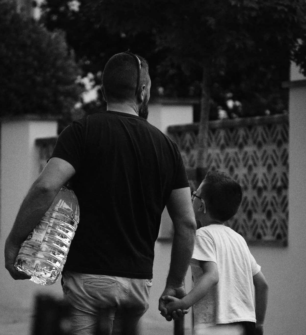 Photo en noir et blanc d'un père et de son jeune fils marchant main dans la main dans la rue, vus de dos, un moment simple du quotidien partagé.