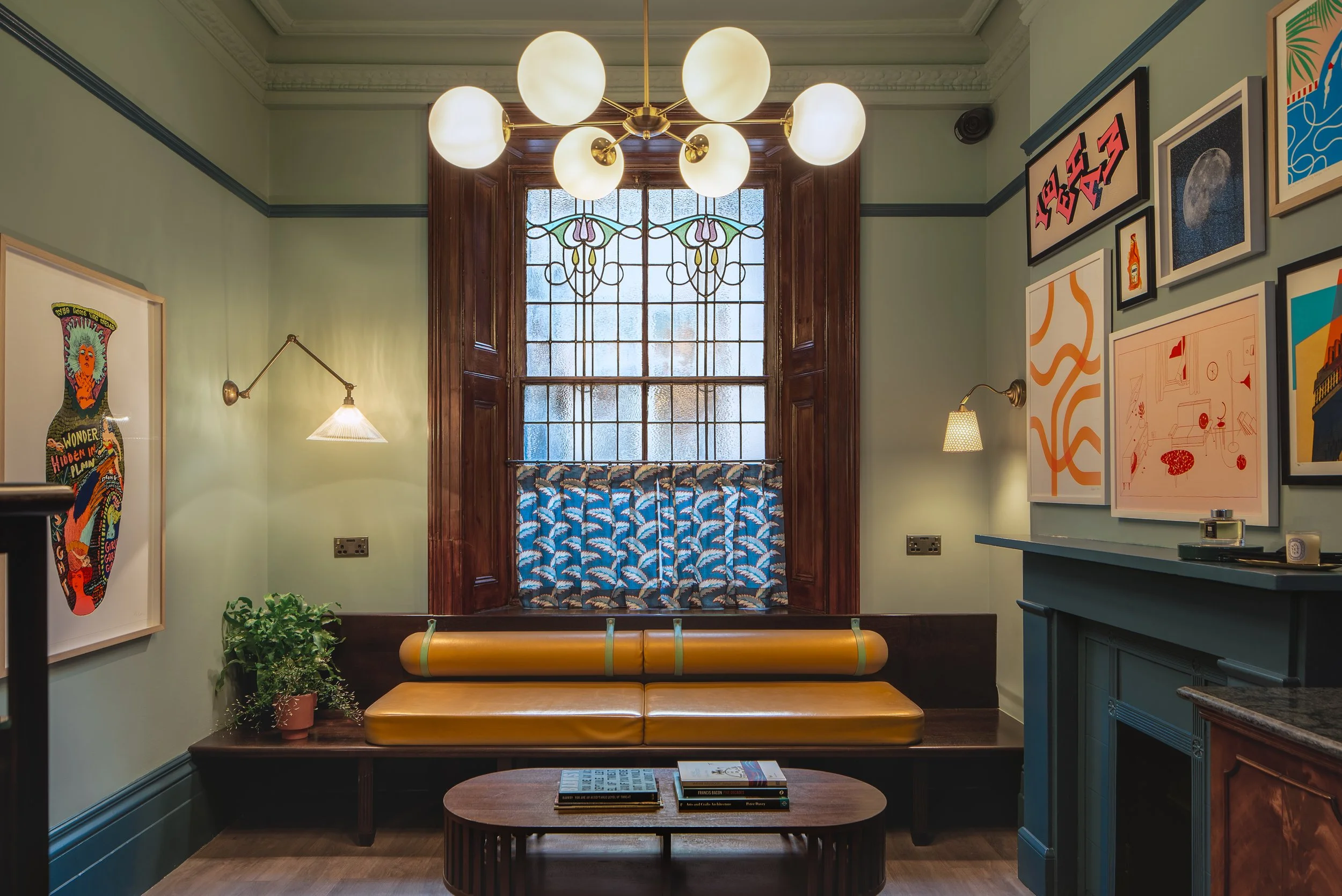 Interior of a cozy living room with a large stained glass window, a yellow leather bench, wall-mounted lamps, and various framed artworks on pastel green walls.
