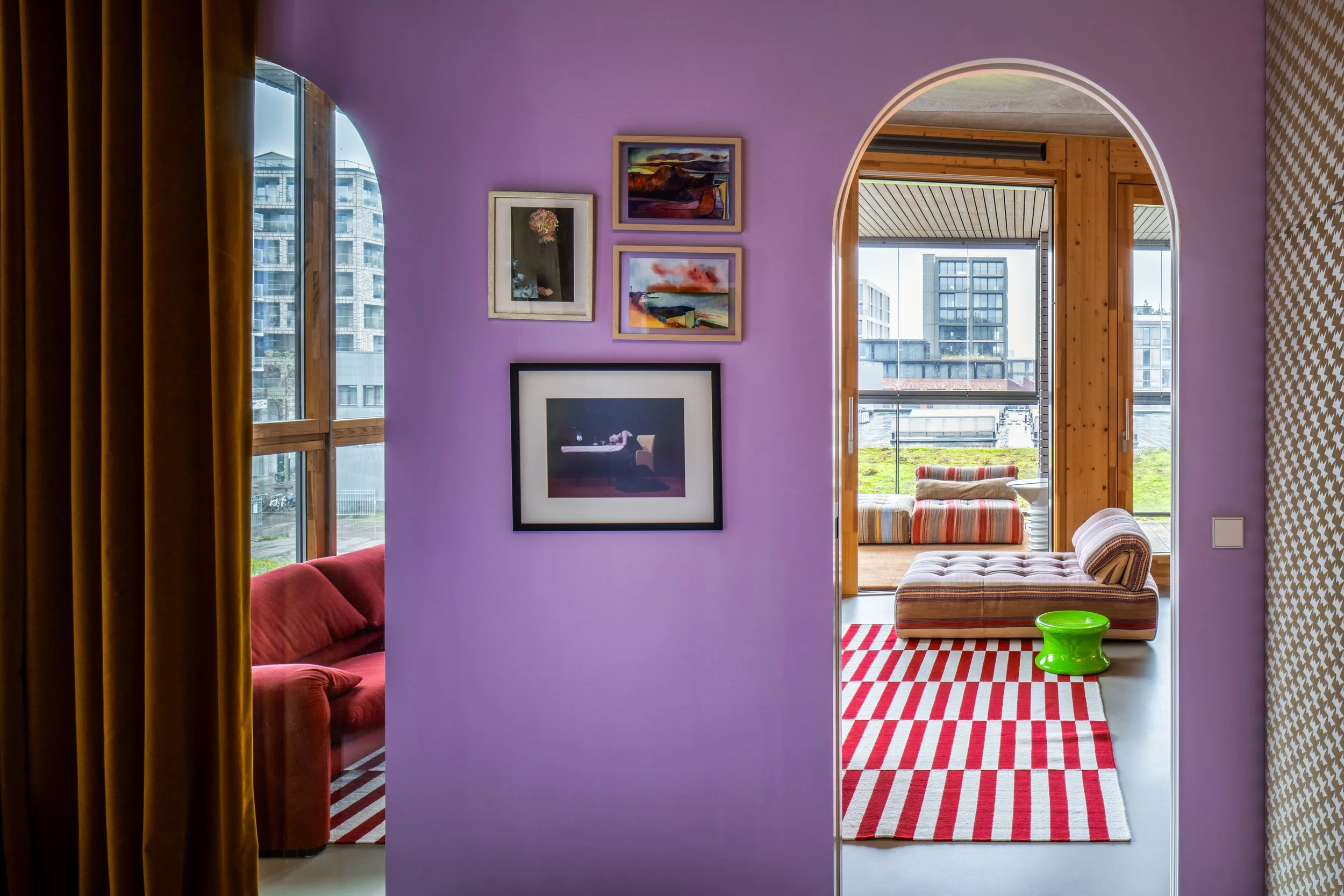 Interior view of an apartment with purple walls, displaying framed artwork, and two rooms with large windows, colorful seating, and patterned rugs.