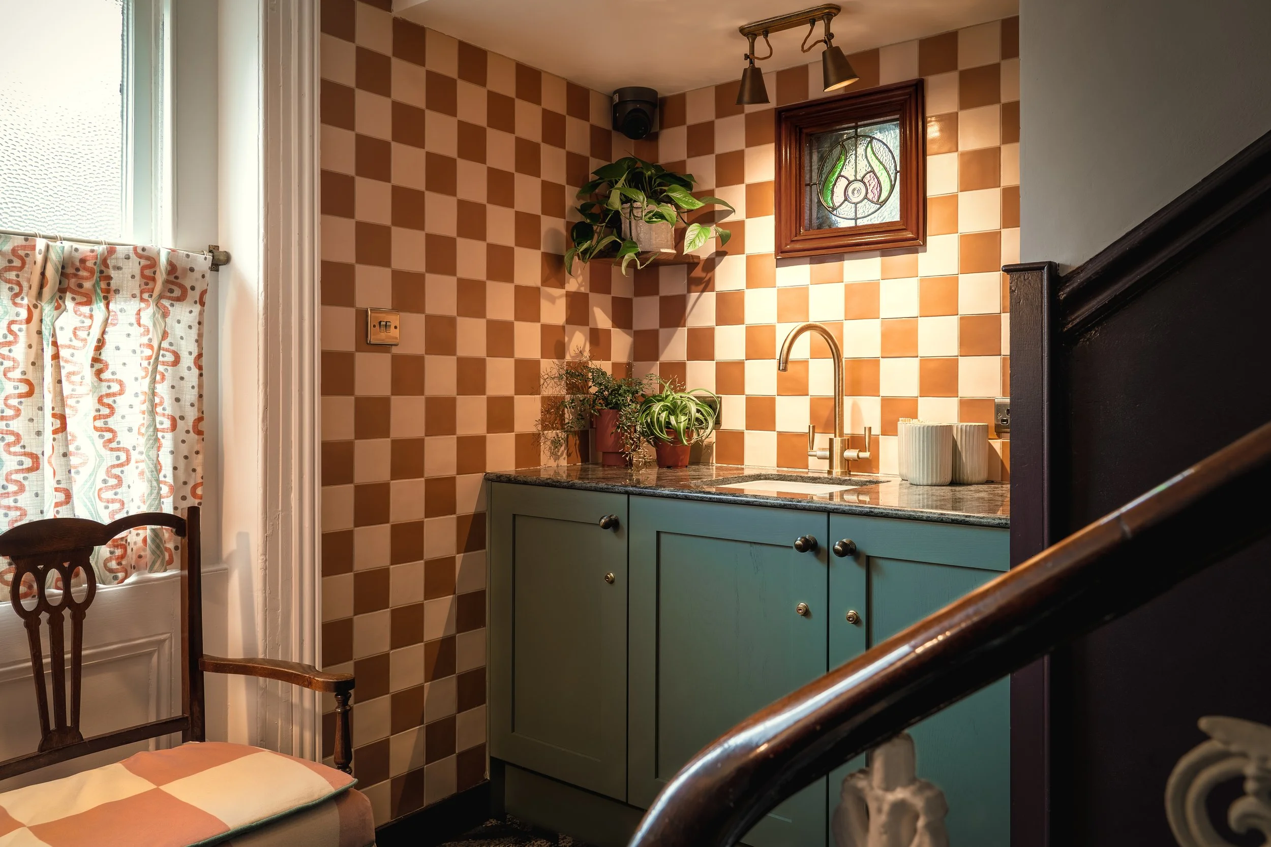 Cozy vintage kitchen corner with a checkered orange and beige backsplash, a small stained glass window, a blue cabinet with a granite countertop, potted plants, and a brass faucet. Part of a wooden chair and a window with patterned curtains are also 