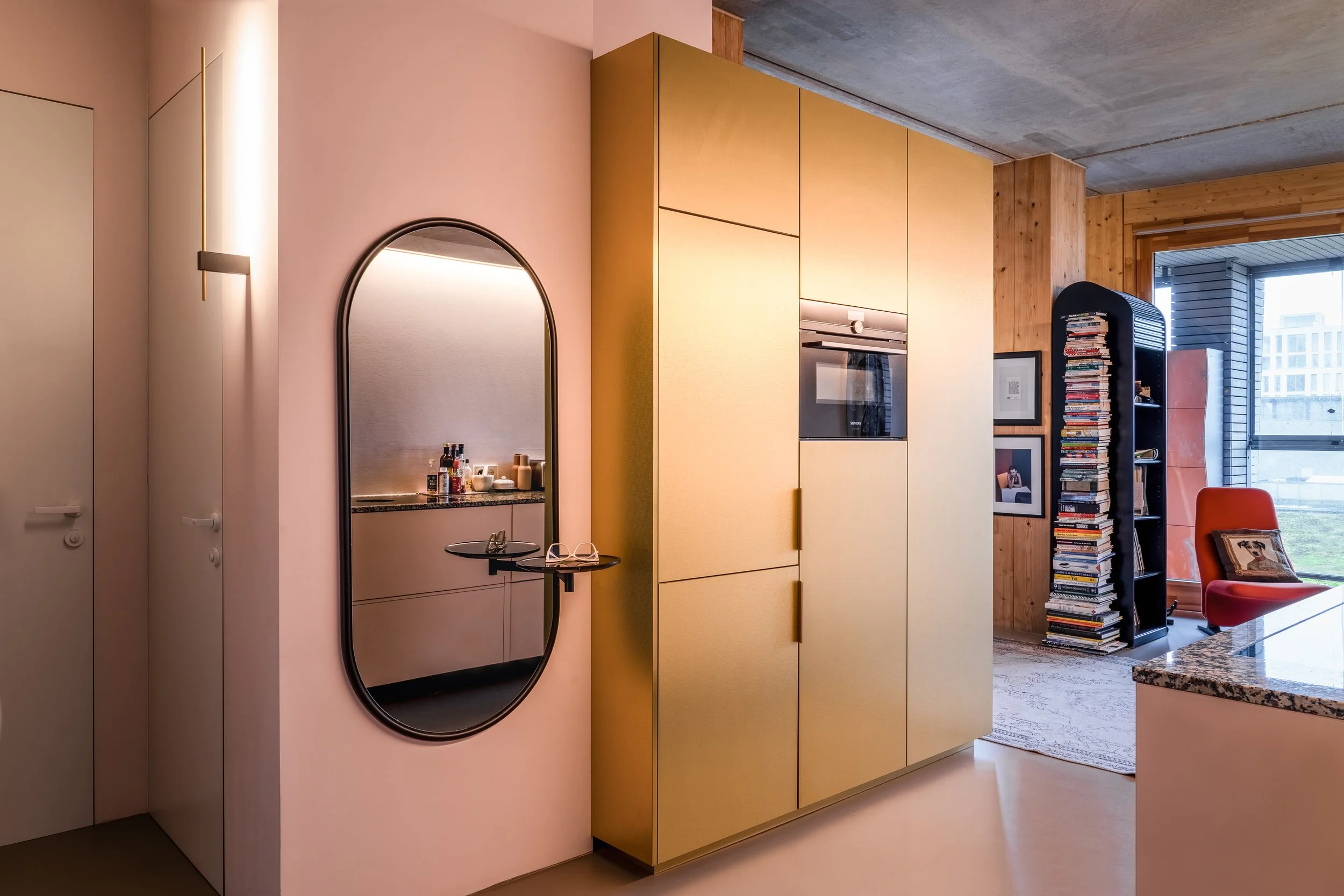 Interior view of a modern apartment with a pink wall, large mirror, gold huede kitchen cabinetry, and a living area with a bookshelf filled with books and a red chair near a large window.