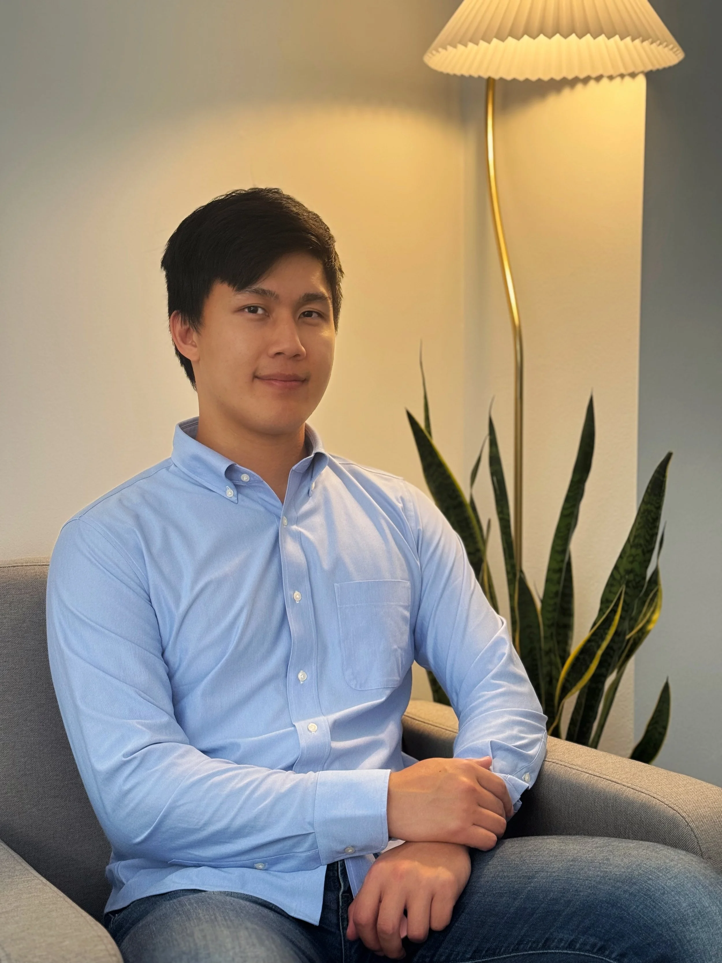 A man with short black hair wearing a light blue button-up shirt, standing in a living room with bookshelves, a lamp, and a window in the background. This is Alex, a psychologist.