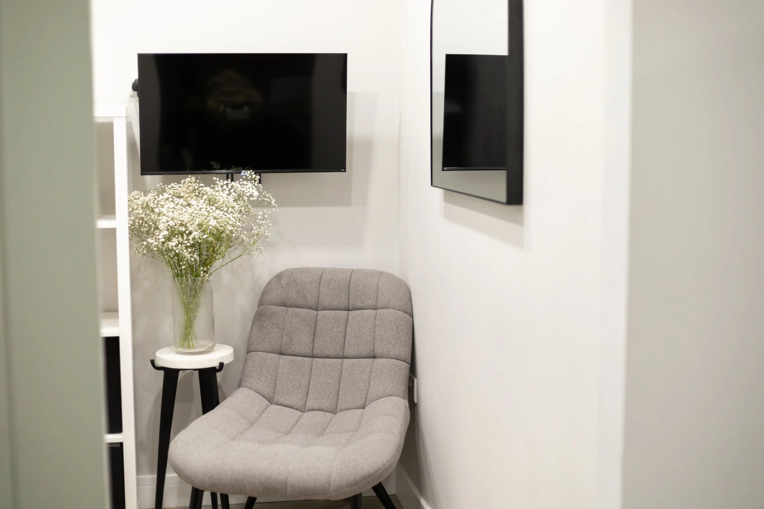 Small cozy corner with a gray upholstered chair, a white side table with a vase of white baby's breath flowers, two wall-mounted flat-screen TVs, and white walls.