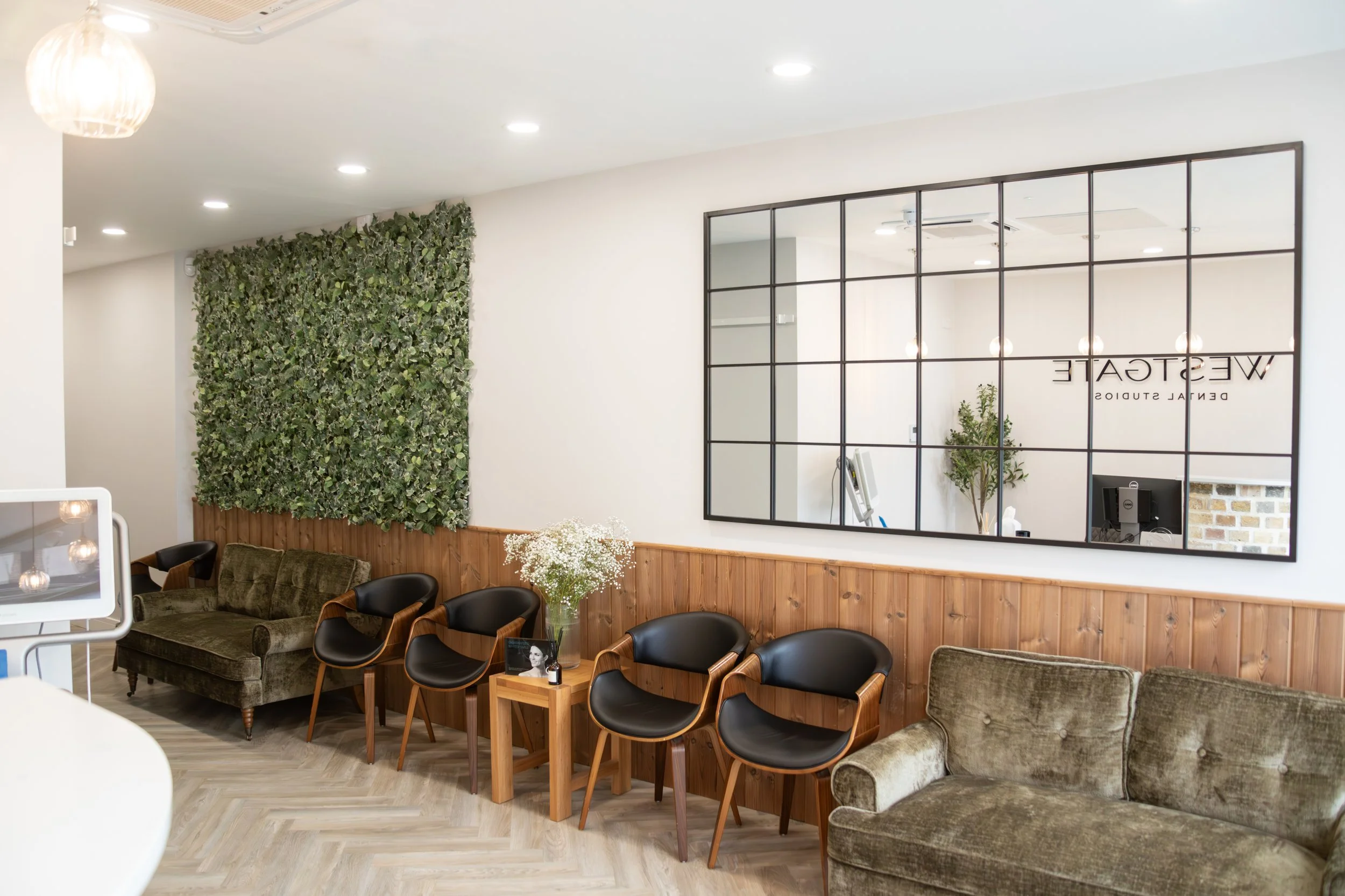 Decorative interior of a waiting area with green velvet sofas, black and wood chairs, a wooden side table with flowers, a large wall mirror, and a green plant, at a wellness or dental studio.