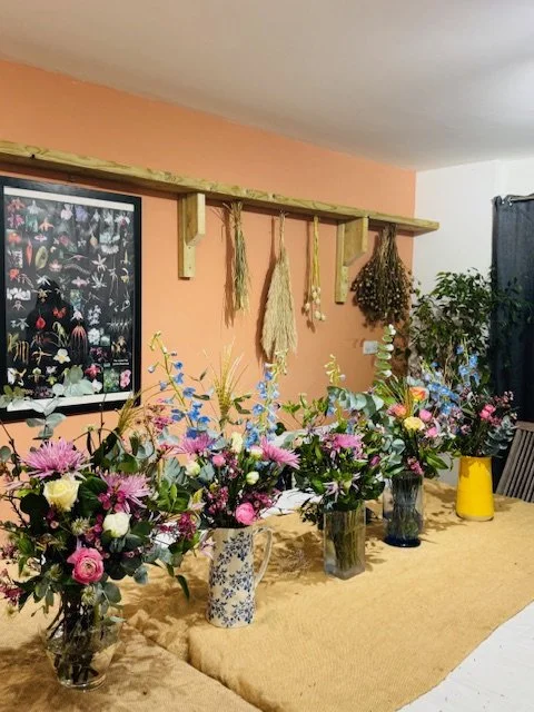 Multiple floral arrangements in various vases and jars on a table, with a pink wall decorated with hanging dried herbs and a picture frame on the left.