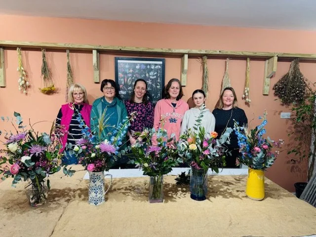 Six women standing behind a long table with colorful floral arrangements in vases, in a room with pink walls and hanging dried flowers.
