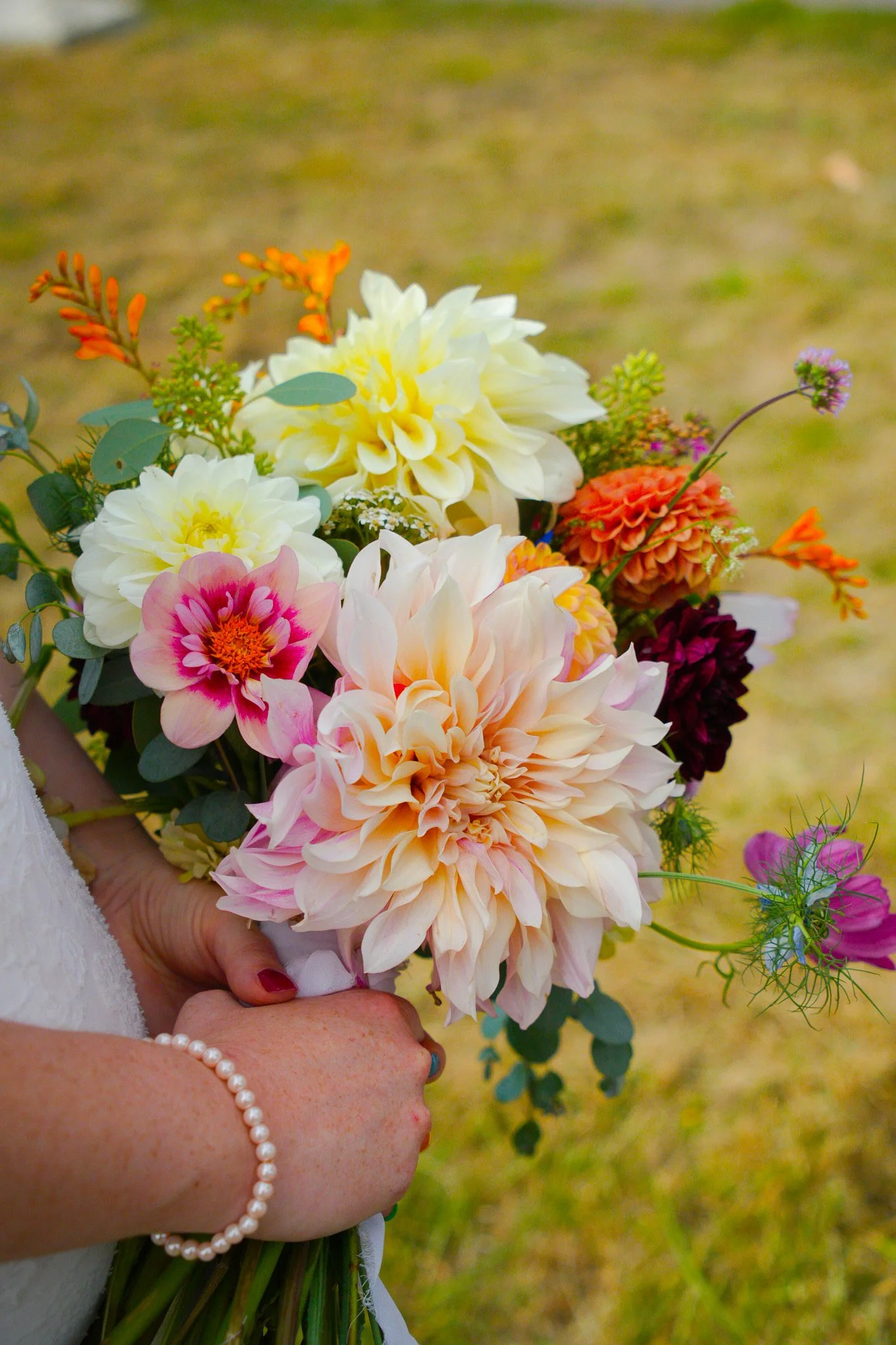A person holding a colorful bouquet of flowers including dahlias, roses, and other blooms, with a blurred background of grass.