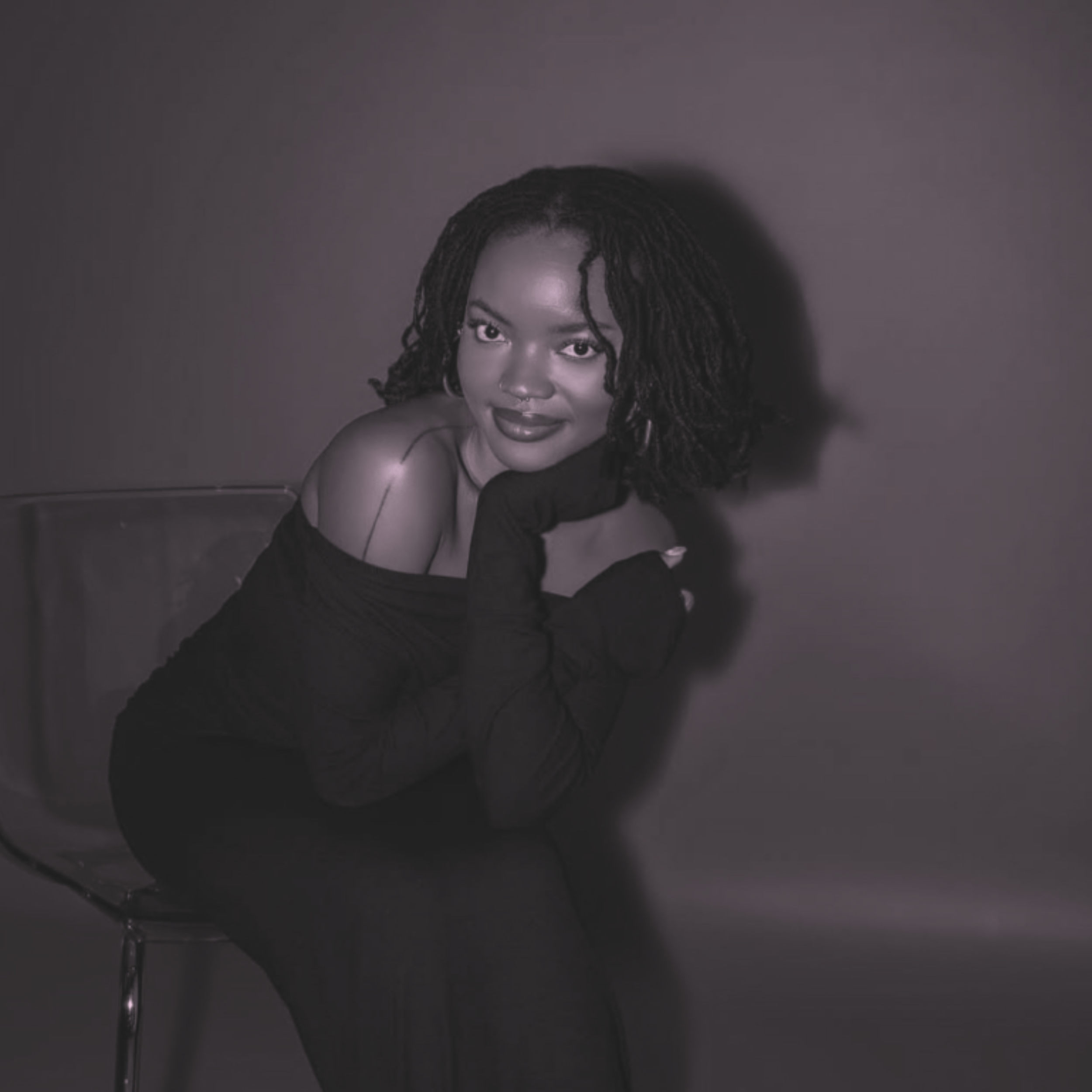 A woman with dark hair styled in twists, makeup, and hoop earrings, wearing an off-shoulder black top, poses against a gray background in a professional portrait setting.