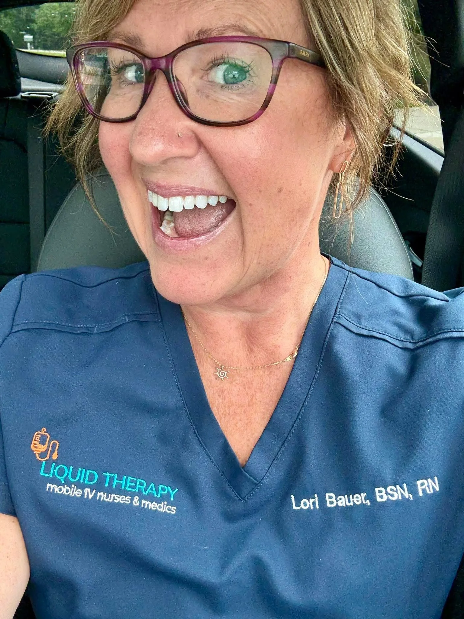 Close-up of a woman with light brown hair, wearing glasses, smiling, and dressed in a blue shirt with embroidered text about liquid therapy, mobile IV nurses, and her name Lori Bauer, BSN, RN.
