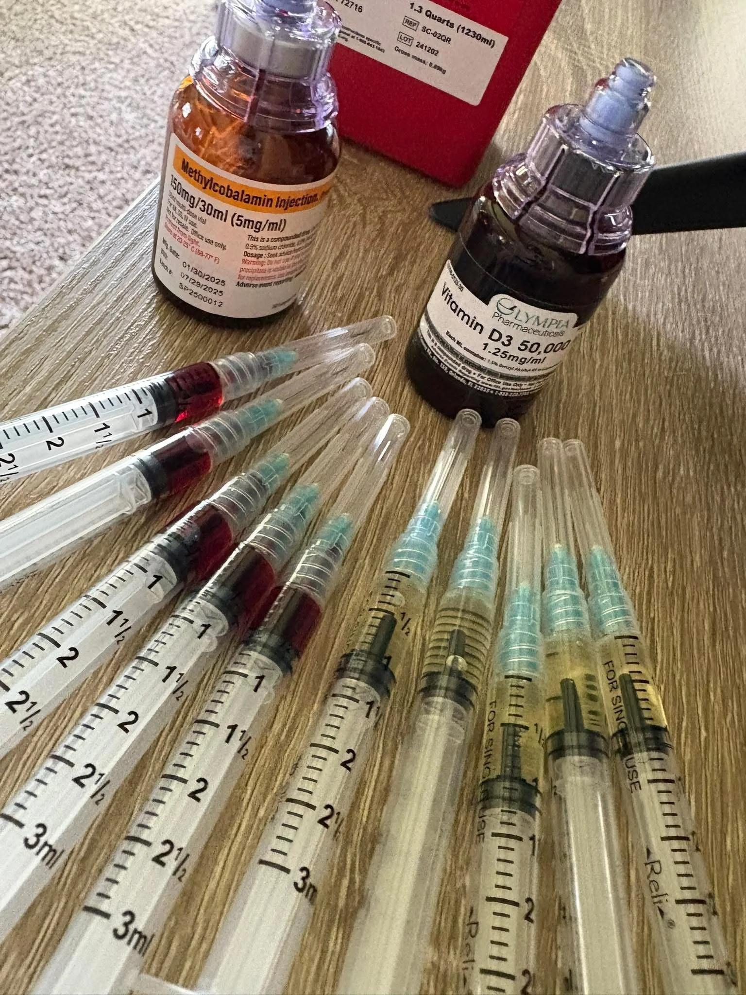 Medical supplies including multiple syringes filled with red liquid, bottles of Methyldobolamin and Vitamin D3, and a red box in the background, all placed on a wooden surface.