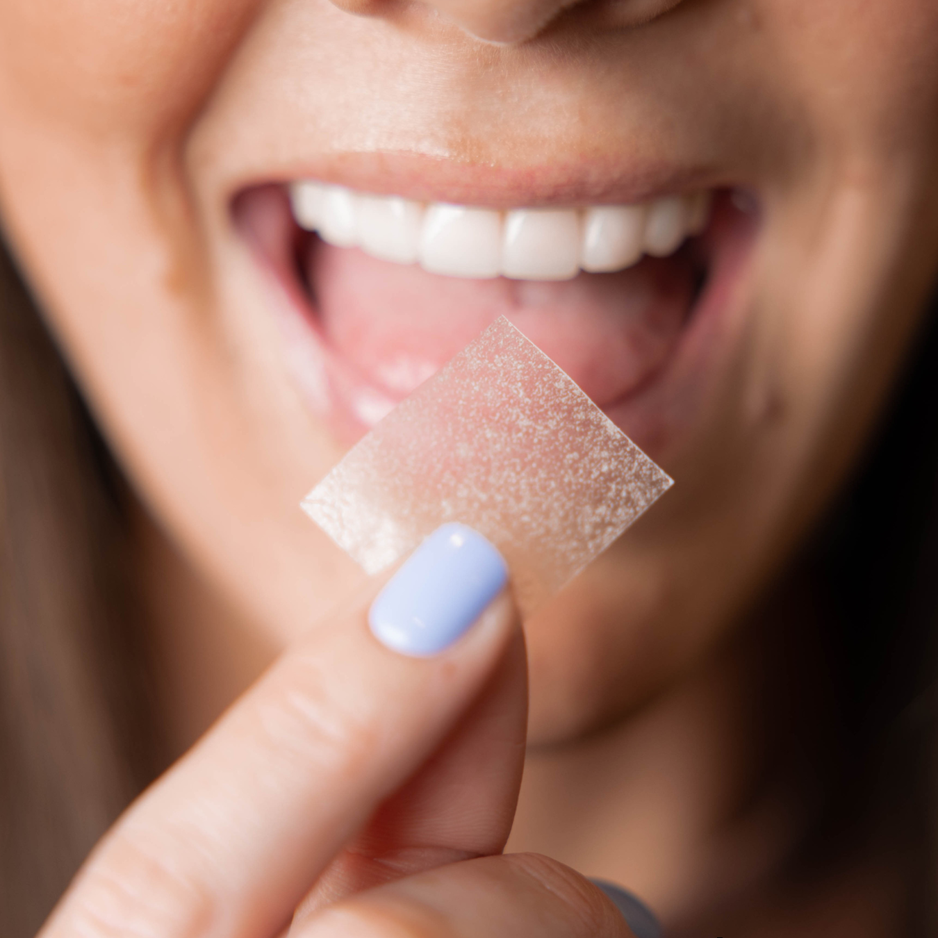 Close-up of a woman with a big smile holding a strip of teeth whitening wax or strip near her open mouth.