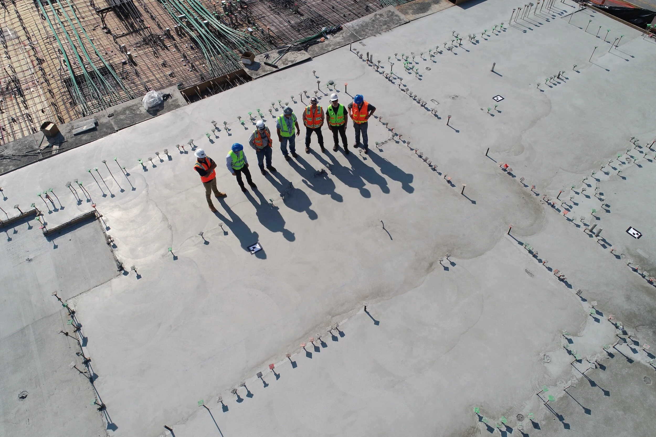 A group of construction workers wearing safety gear standing on a concrete building floor, casting long shadows, with various construction materials and rebar in the background.