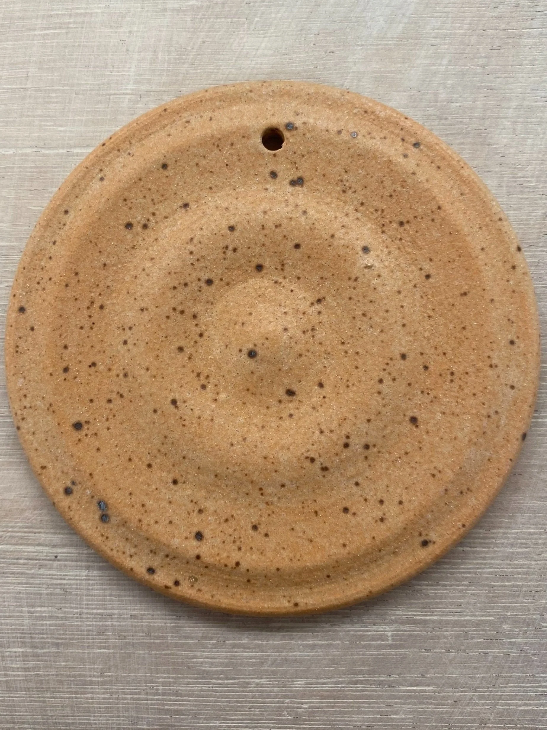 A round, beige, speckled ceramic plate with a small hole at the top, placed on a light wood surface.
