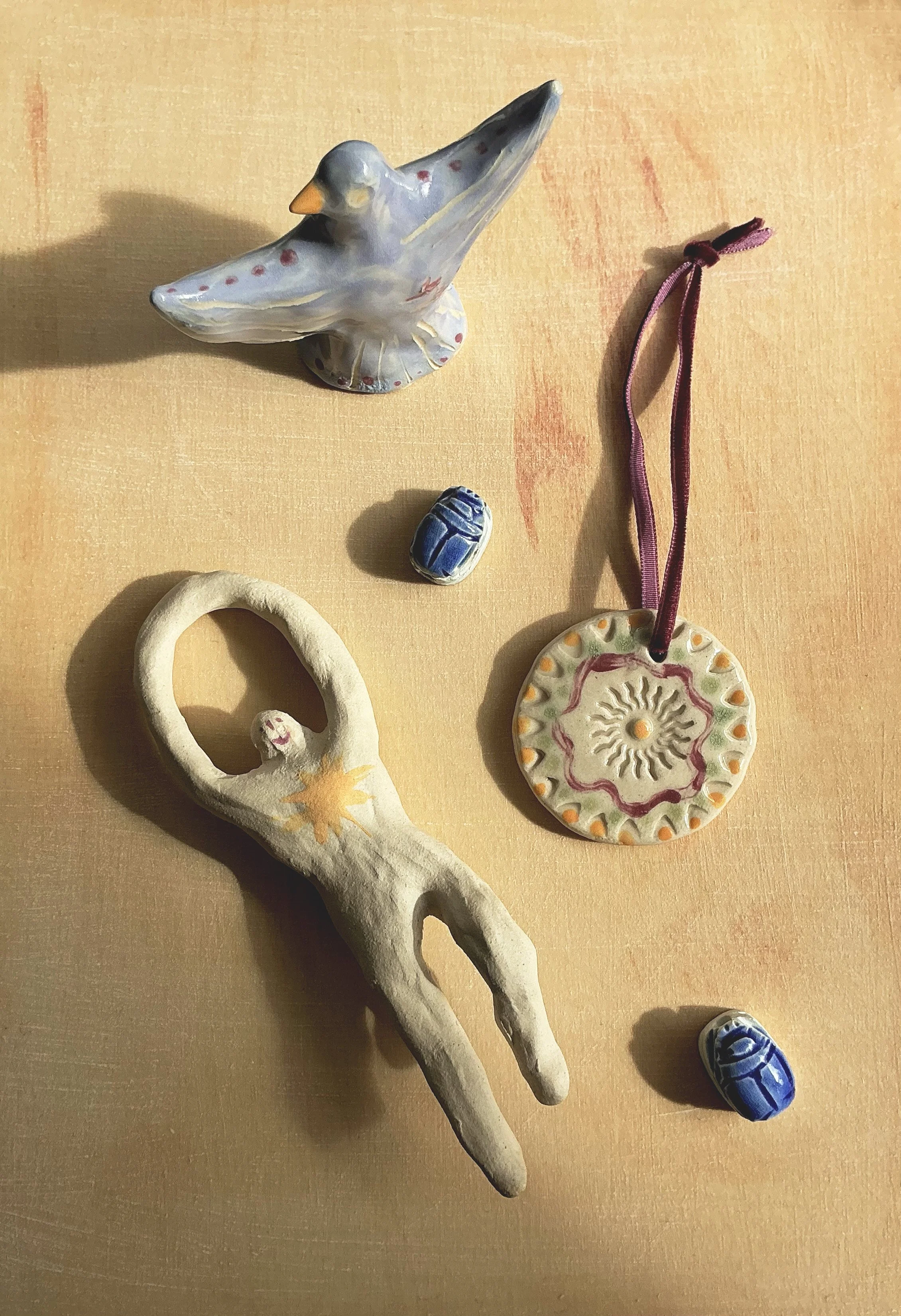 Collection of handcrafted ceramic and clay objects on a wooden surface, including a bird figurine, a circular pendant with a purple ribbon, a face-shaped piece, and several small ear-shaped sculptures.