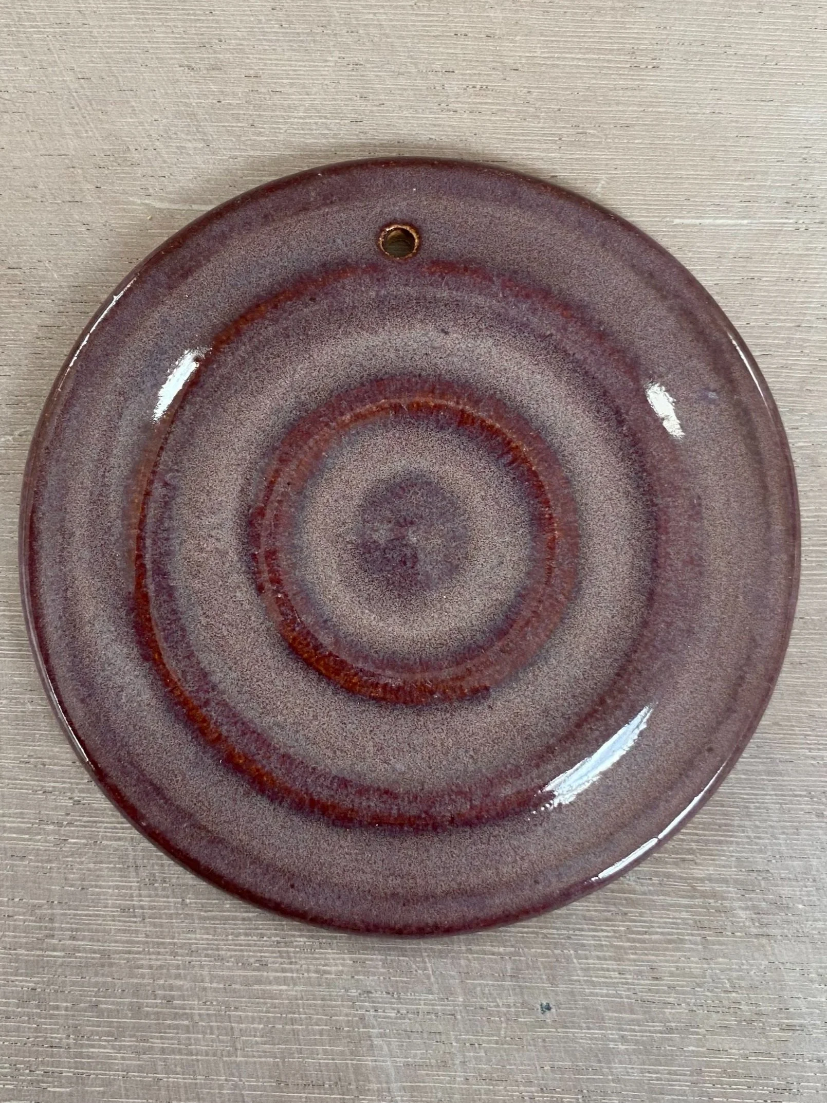 A round ceramic plate with a spiral pattern in shades of brown and purple, on a light wood surface, with a small hole at the top edge.