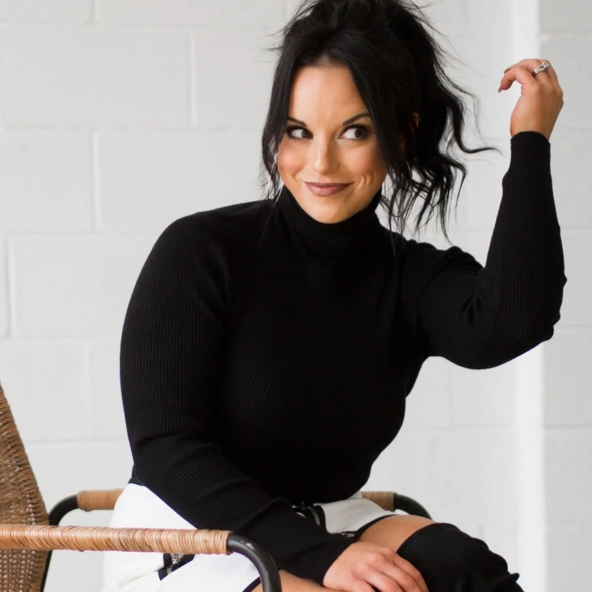 photo of mindy thomas in black turtle neck sweatear, white and black skirt, sitting in chair.