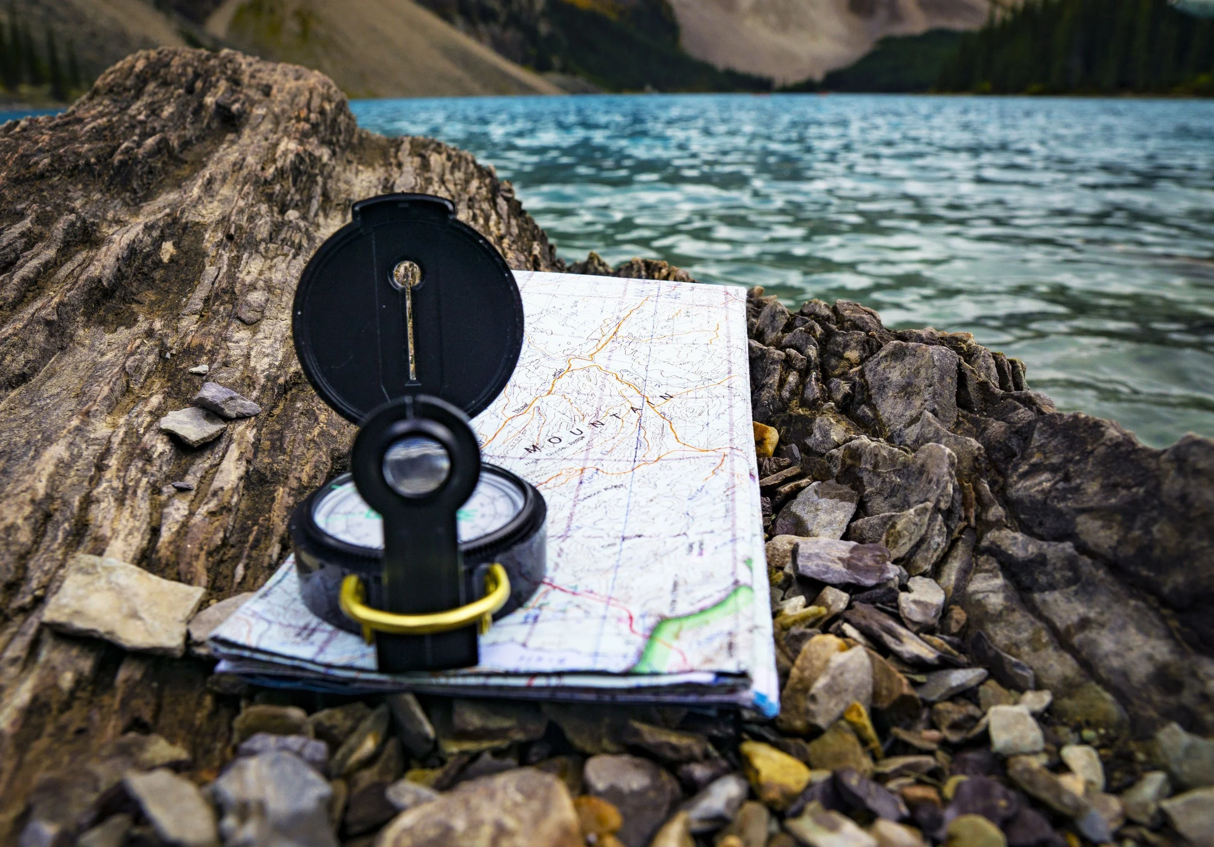 A compass and a trail map resting on rocks beside a mountain lake, with mountains and a forests in the background.