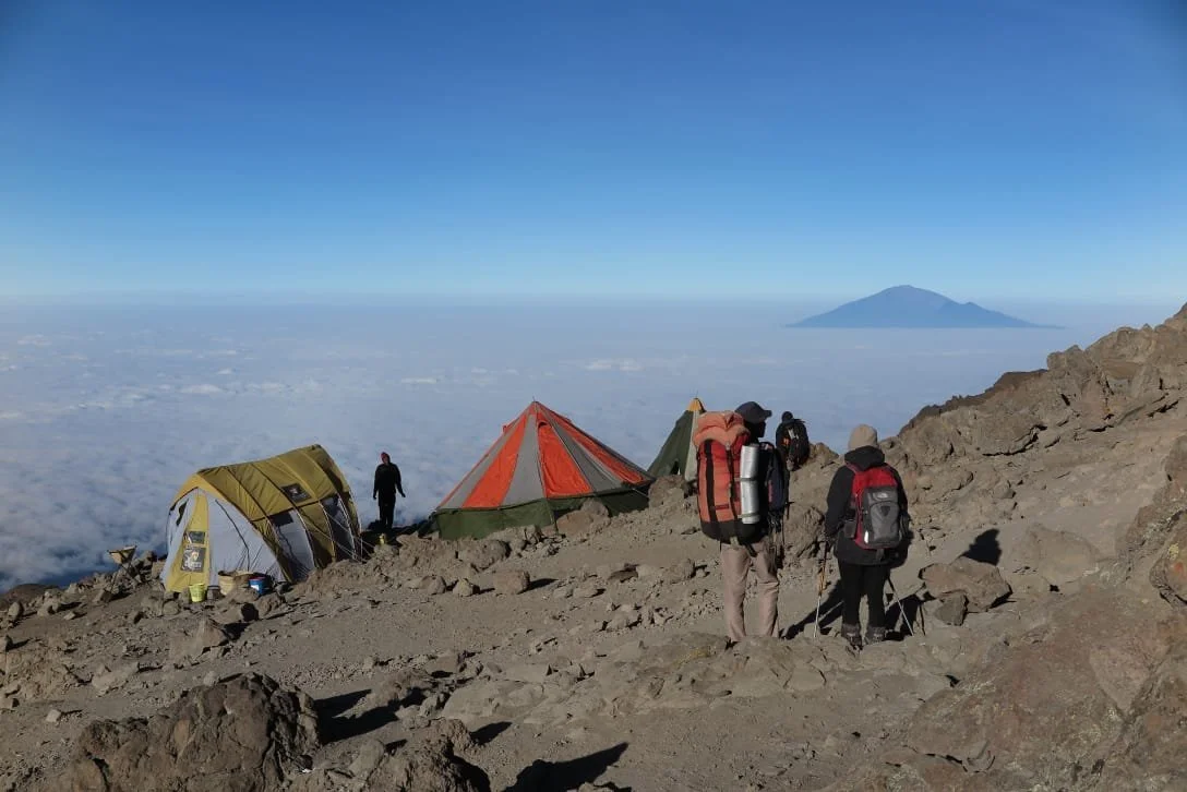 Group of hikers with backpacks camping near tents on a mountain top above the clouds, with a distant island visible on the horizon under a clear blue sky.