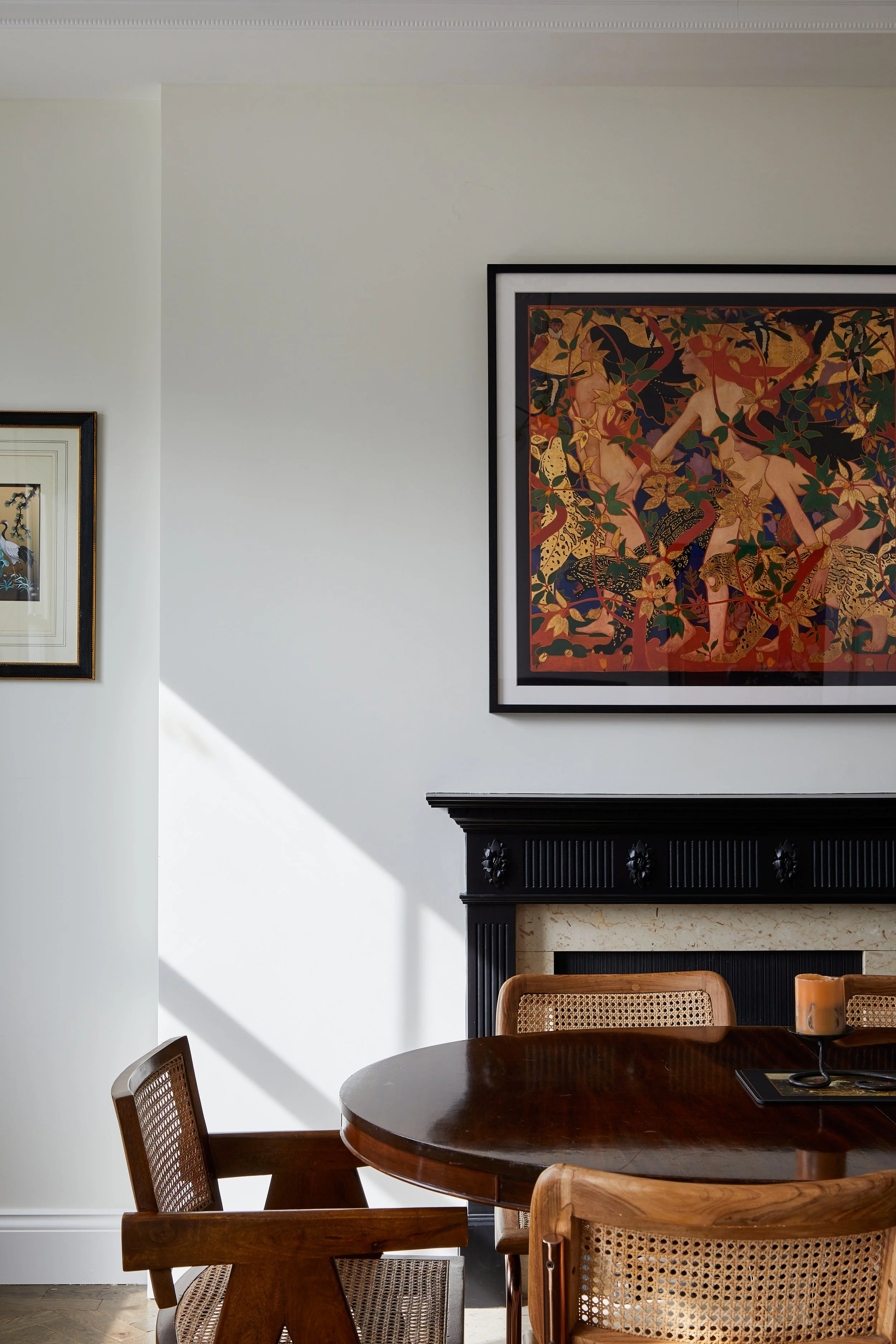 A dining room in a Georgian townhouse in Pimlico features a round wooden table and chairs. Above a black fireplace, colorful framed artworks adorn the walls. Sunlight casts shadows across the white walls and floor, highlighting the elegance of this t