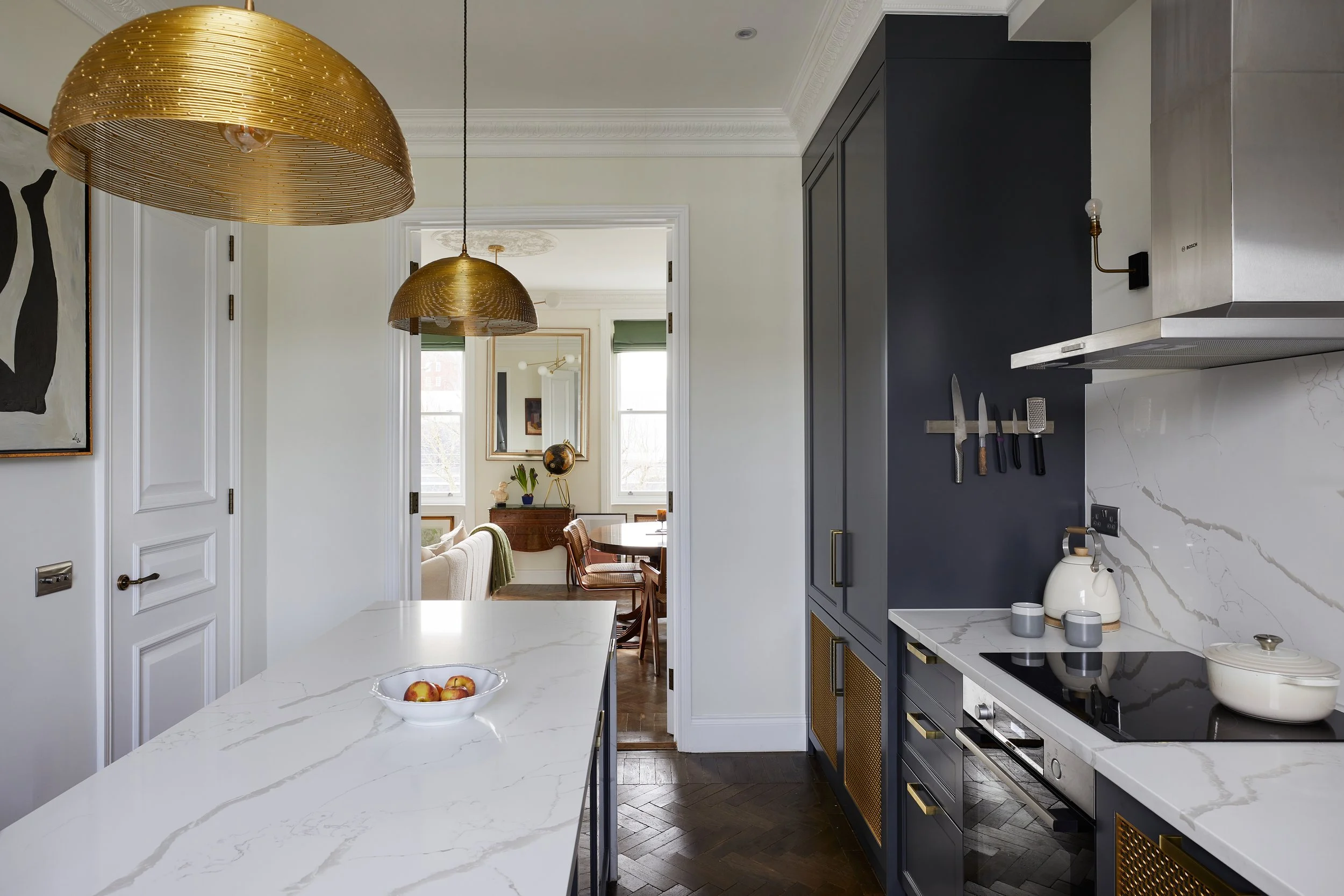 A modern kitchen with a marble island counter, gold pendant lights, and a fruit bowl. Dark cabinetry contrasts with light walls. The kitchen leads to a cozy dining room with a view of green curtains and natural light.